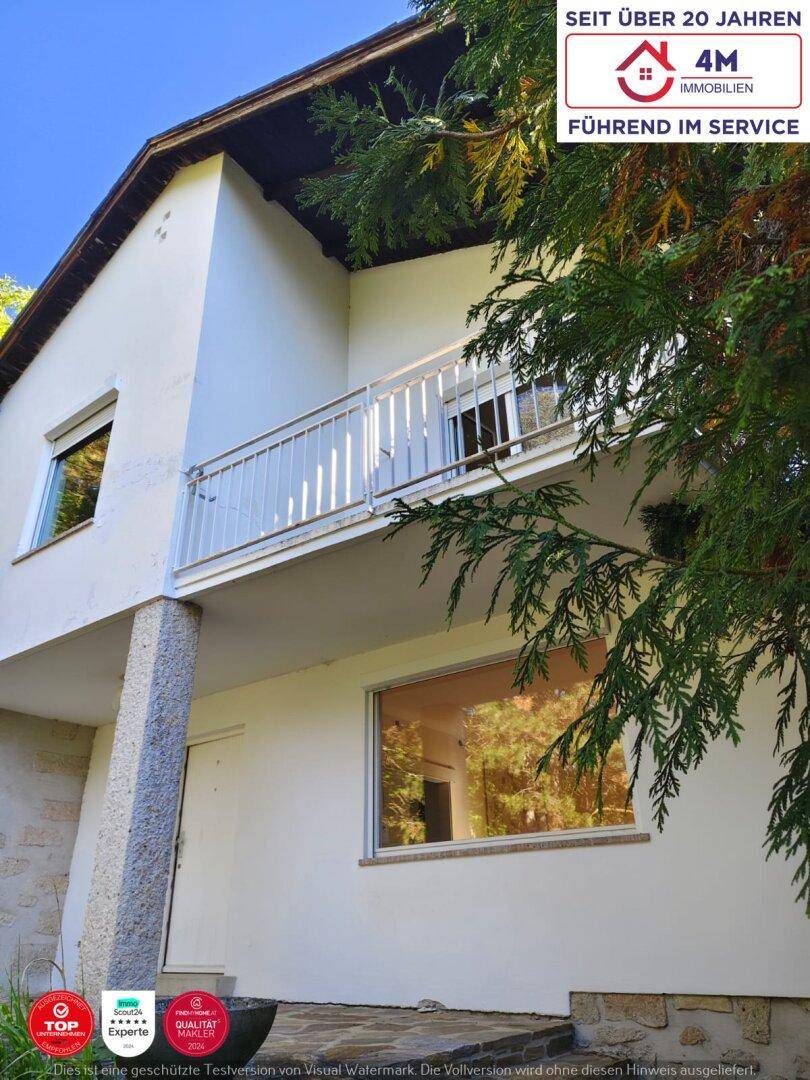 Weißes Haus mit Balkon und Fenstern, umgeben von grünen Bäumen.