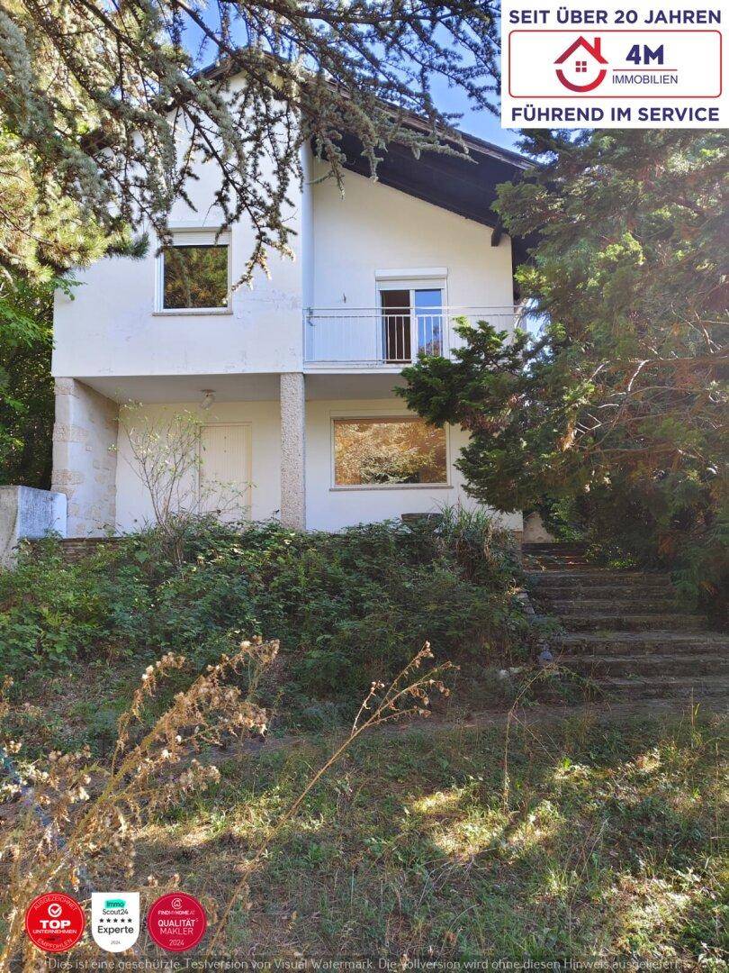 Hausfassade mit Balkon und einem verwilderten Gartenbereich mit Steintreppe.