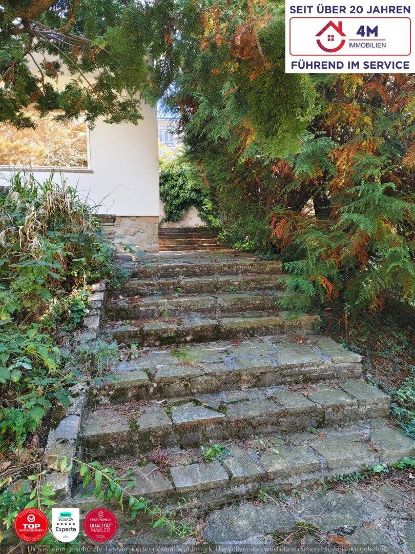 Ein steinerner Gartenweg führt durch üppiges Grün und Bäume zu einem Gebäude.