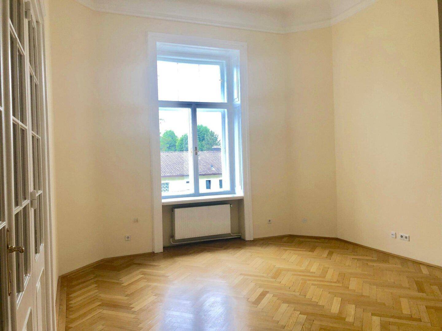 Helles, leeres Zimmer mit Fischgrätparkett, hohen Decken und großem Fenster.