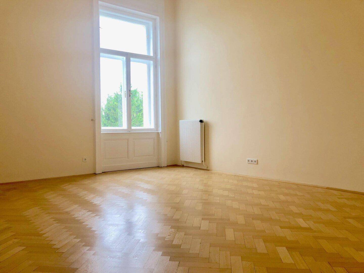 Lichtdurchflutetes, leeres Zimmer mit Fischgrätparkett und hoher Fenstertür.