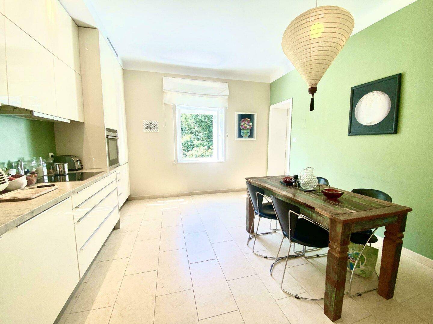 Moderne Küche mit weißen Schränken, einem Holztisch und einer grünen Wand, mit Blick ins Grüne.