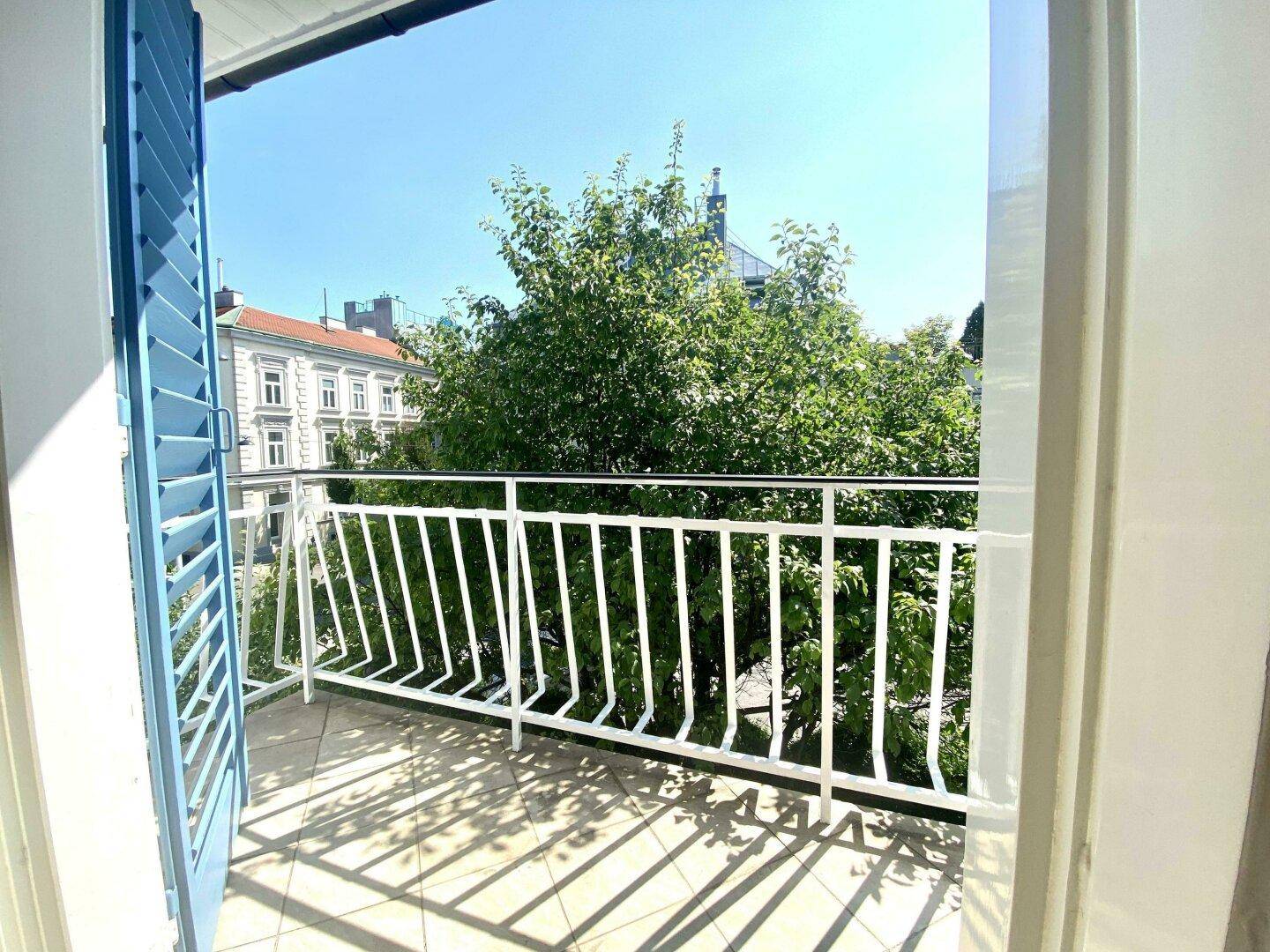 Kleiner Balkon mit weißem Geländer und blauen Fensterläden, umgeben von Bäumen und Stadtgebäuden.