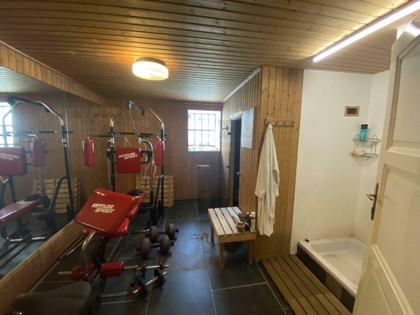 Fitnessraum mit Holzverkleidung, Trainingsgeräten, einer Sauna und einer Duschgelegenheit.