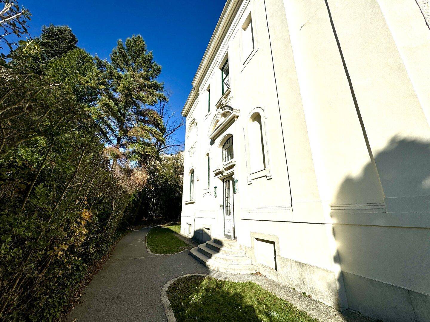 Seitenansicht der Villa mit gepflegtem Gartenweg und hohen Bäumen, umgeben von viel Grün.