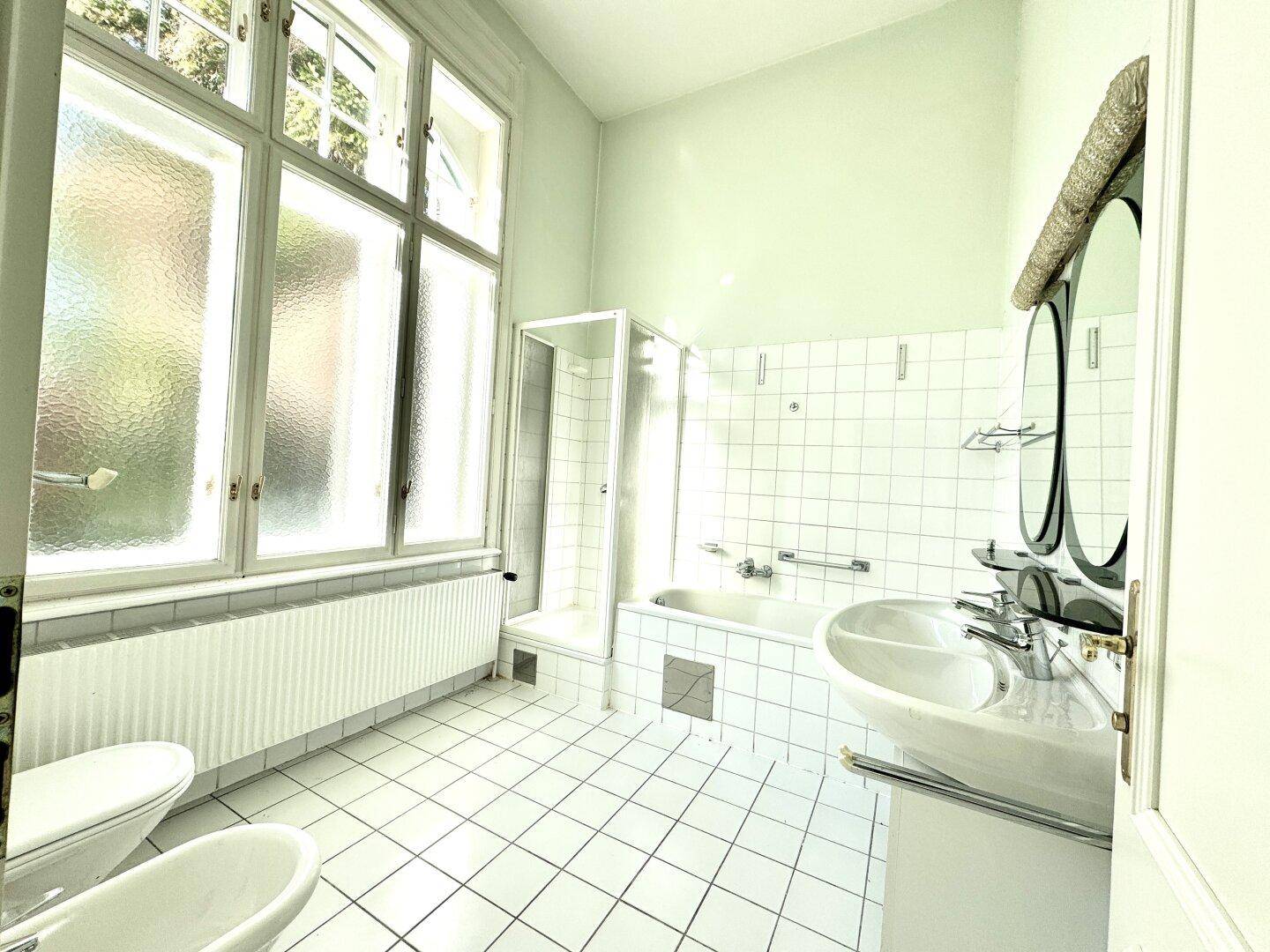 Helles Badezimmer mit weißen Fliesen, Badewanne, Dusche, Bidet und großem Fenster für Tageslicht.