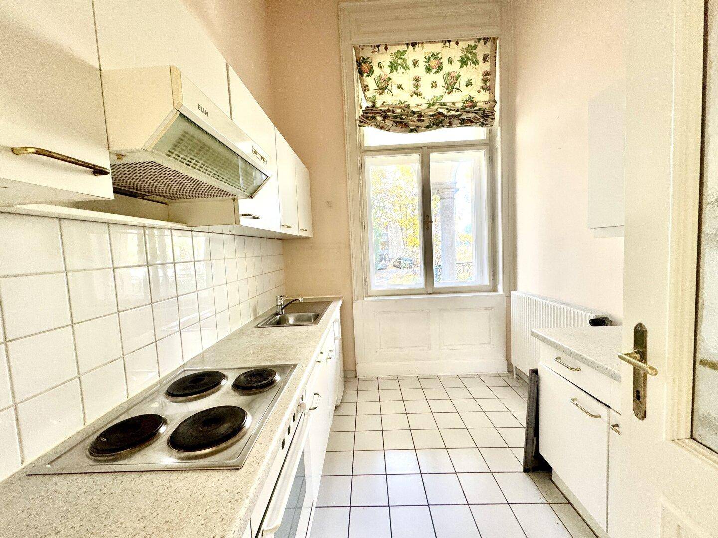 Funktionale Küche mit weißen Schränken, Elektroherd und Fenster mit Blick ins Grüne.