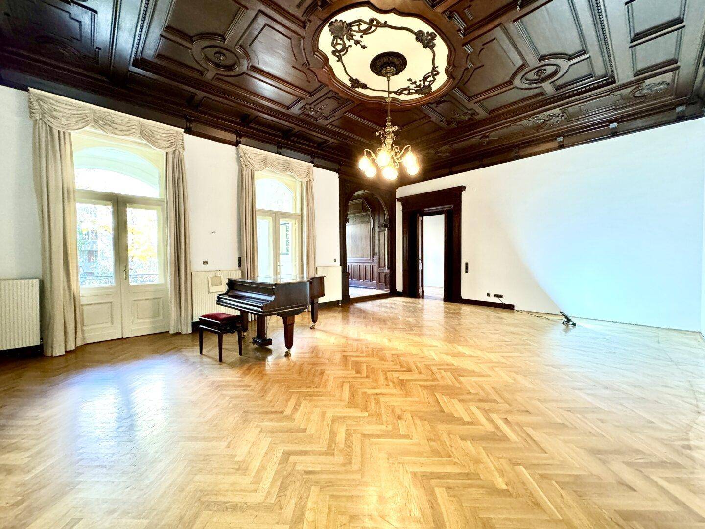 Geräumiger Salon mit beeindruckender Kassettendecke, Kronleuchter und Fischgrätparkett, ideal für Empfänge.