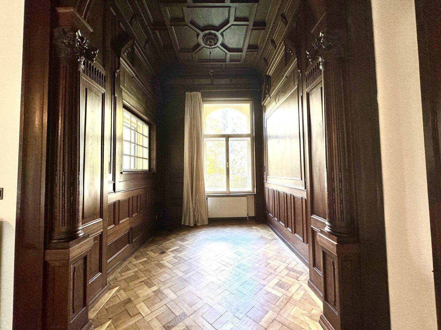 Repräsentativer Flur mit dunkler Holzvertäfelung, Kassettendecke und Fischgrätparkett, lichtdurchflutet.