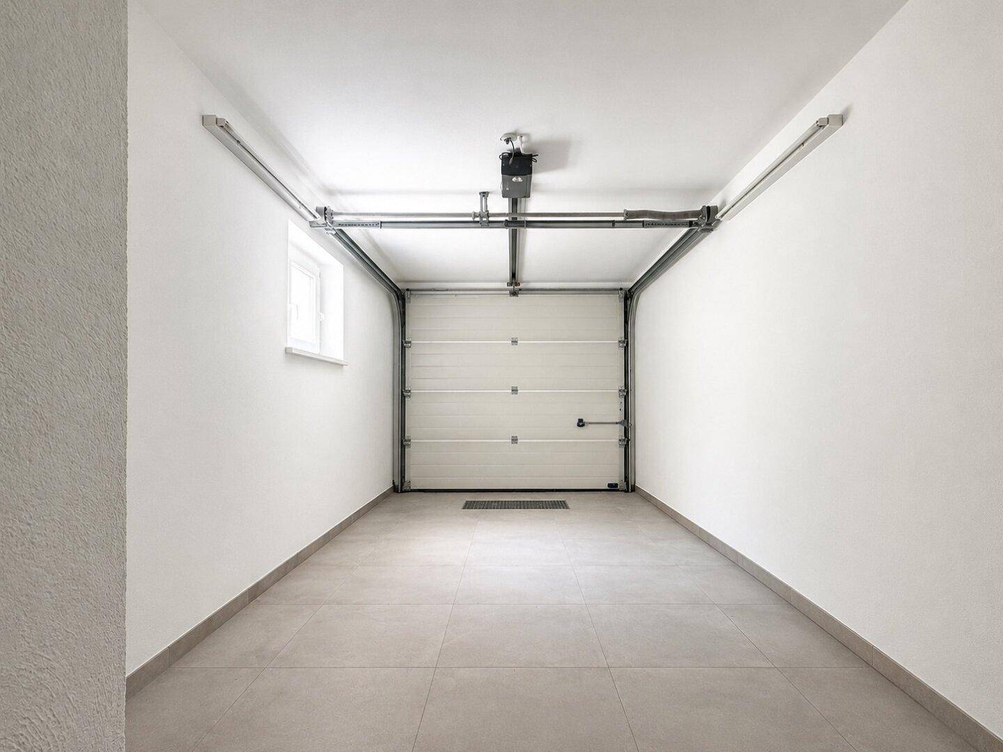 Leere Garage mit automatischem Tor, kleinem Fenster und Fliesenboden.