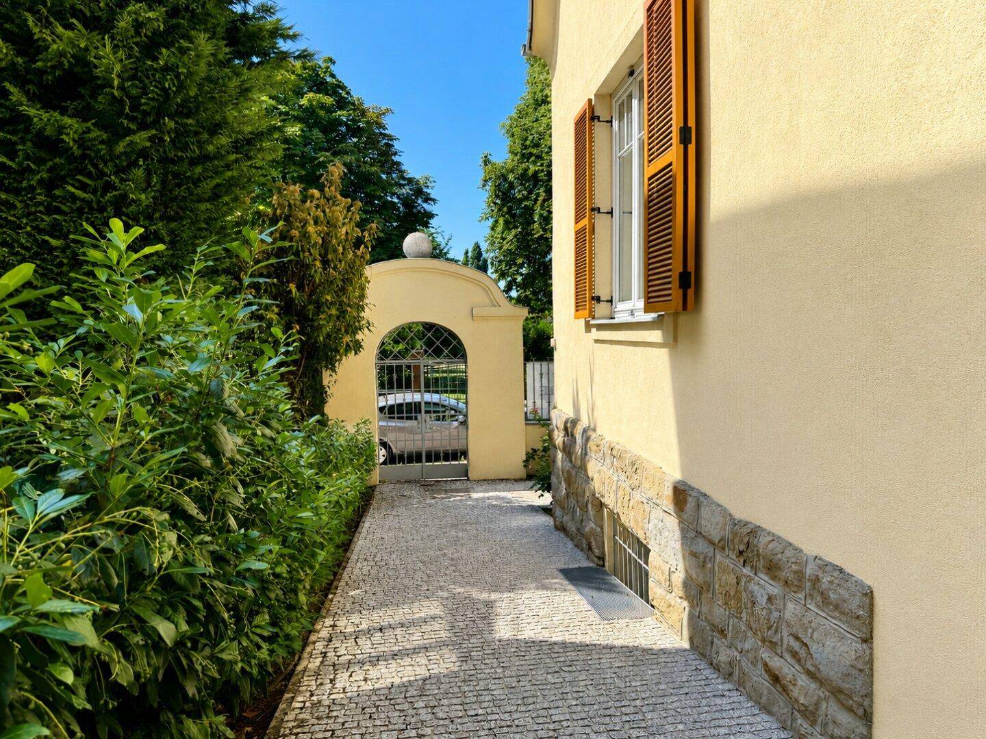 Sonniger Eingangsbereich mit gepflastertem Weg, grüner Hecke und historischem Torbogen.