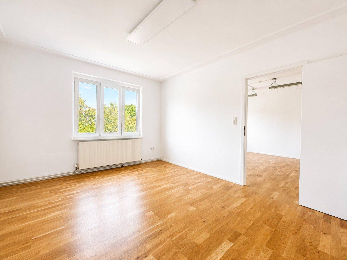 Geräumiger, heller Raum mit Holzboden und Zugang zu einem weiteren Zimmer.