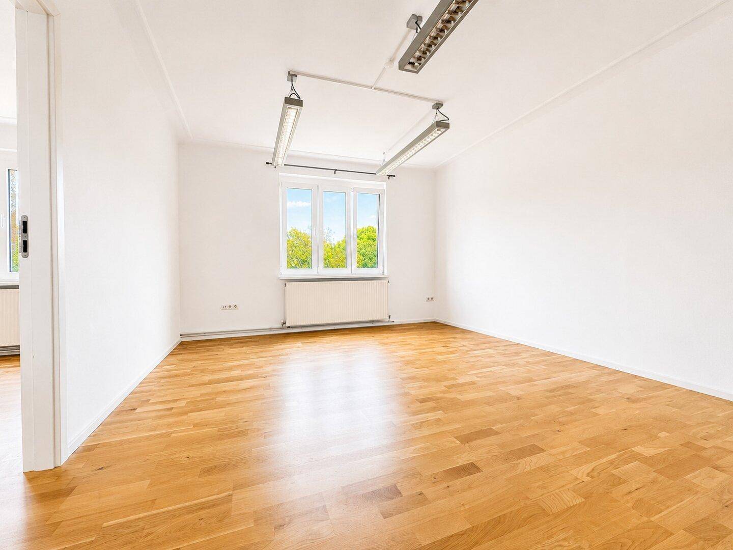 Heller, leerer Raum mit Holzboden, weißen Wänden und großen Fenstern.