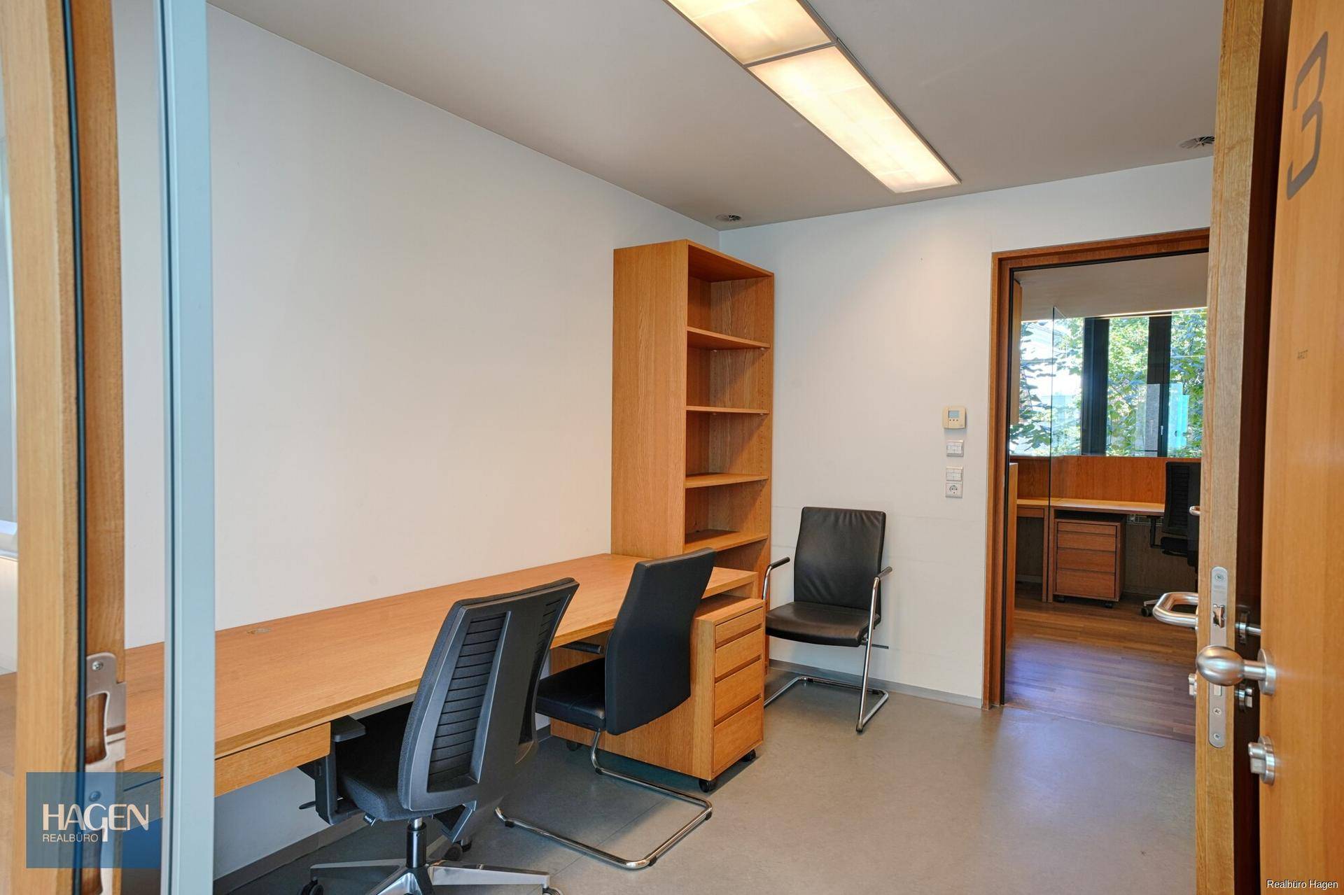 Büro mit Holzmöbeln, zwei Arbeitsplätzen und Blick in einen weiteren Bürobereich.