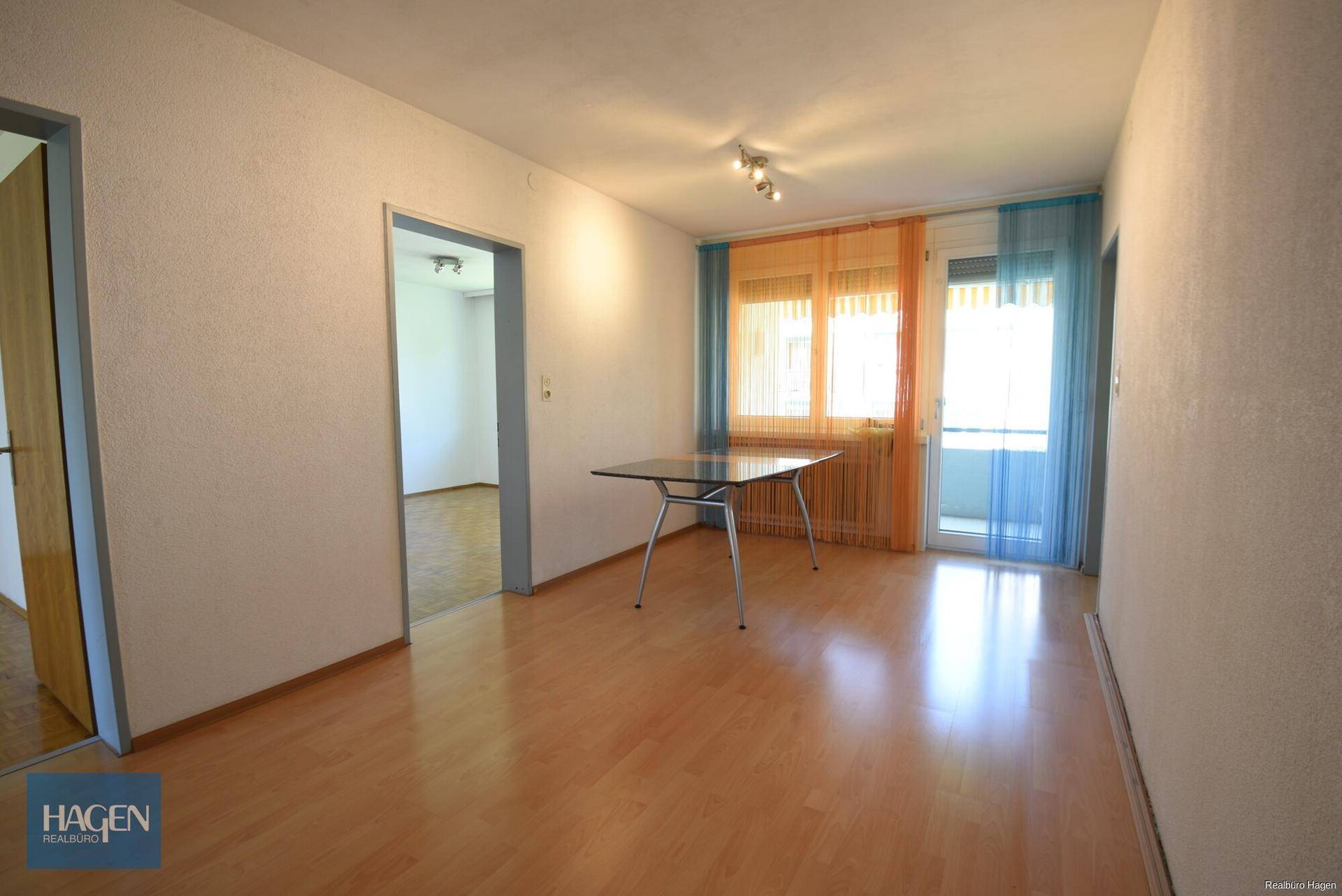 Wohnzimmer mit Laminatboden, Balkonzugang und farbigen Vorhängen.