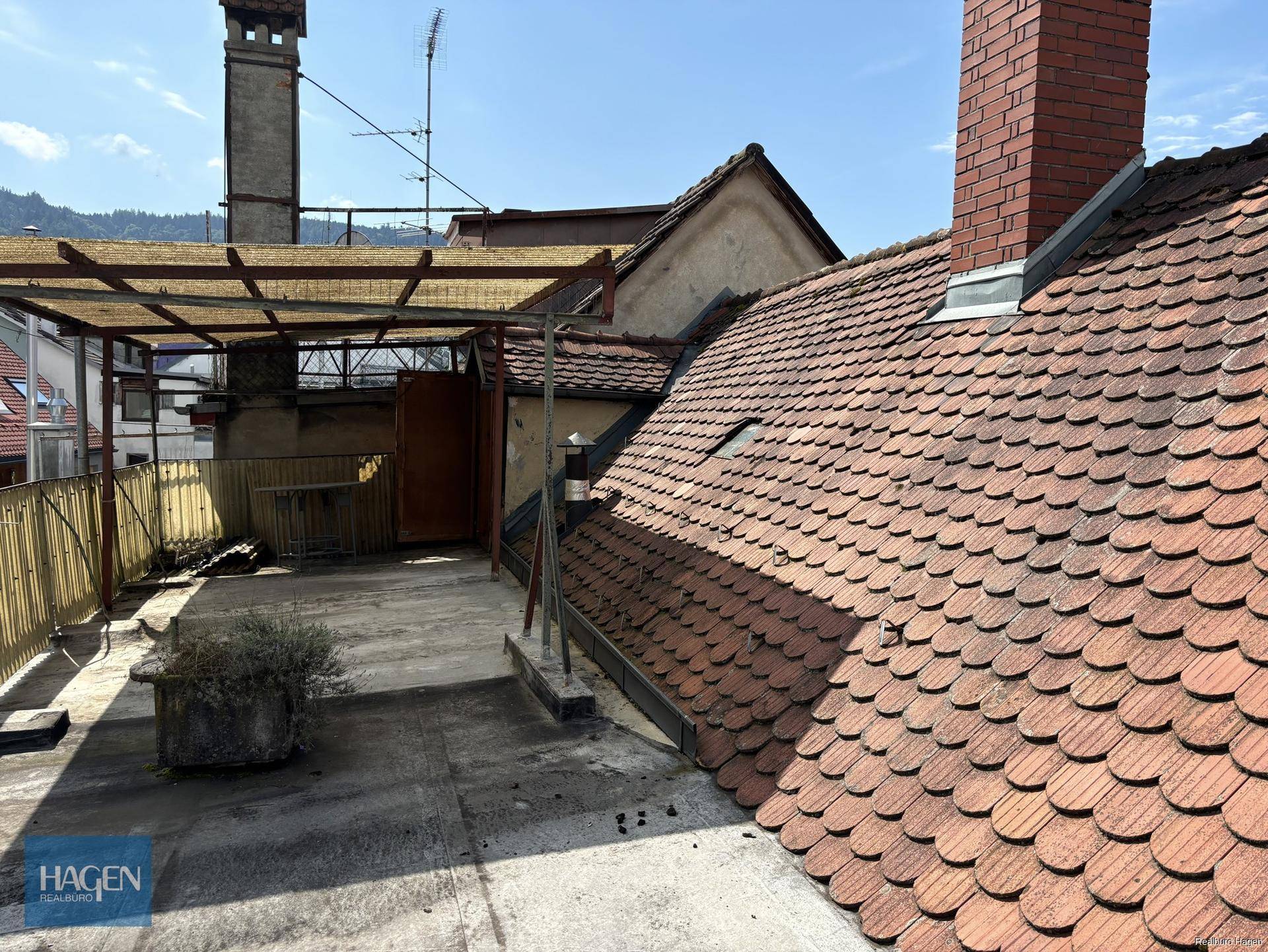Dachterrasse mit Blick auf umliegende Dächer und Schornsteine unter blauem Himmel.