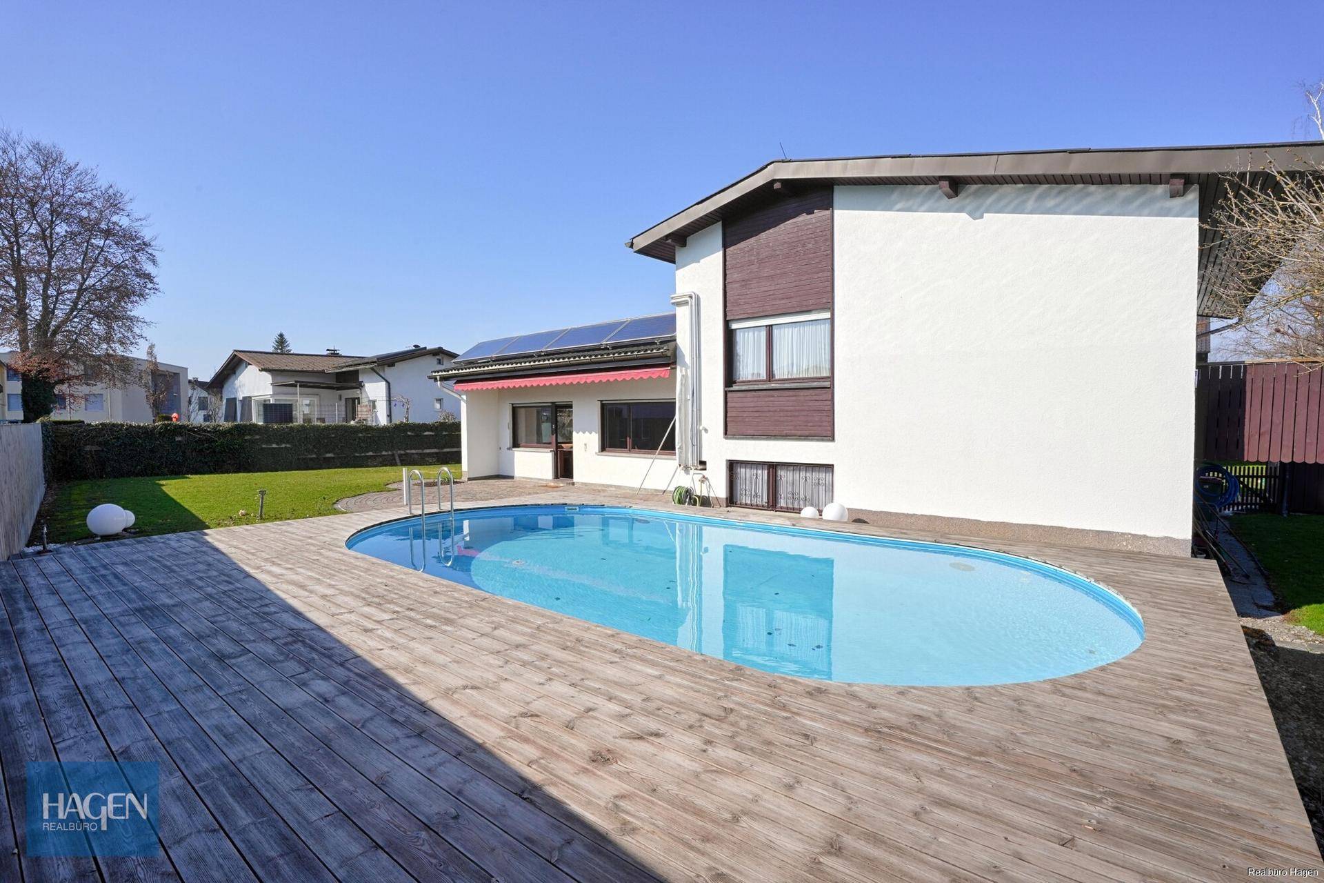 Ovaler Swimmingpool mit Holzdeck und Blick auf das Wohnhaus.