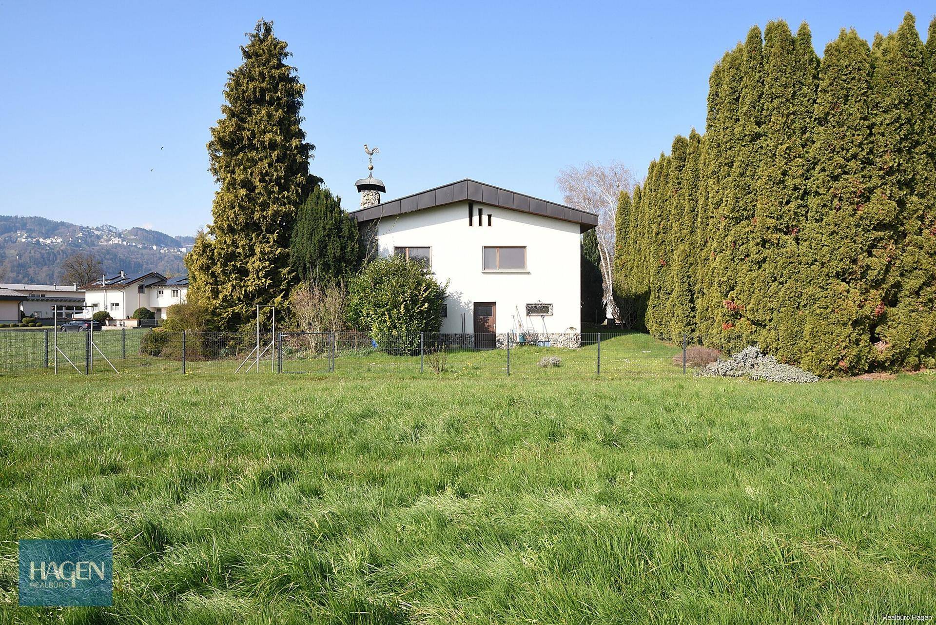 Freistehendes Haus mit weißer Fassade und Satteldach, umgeben von hohen Hecken und Rasenfläche.