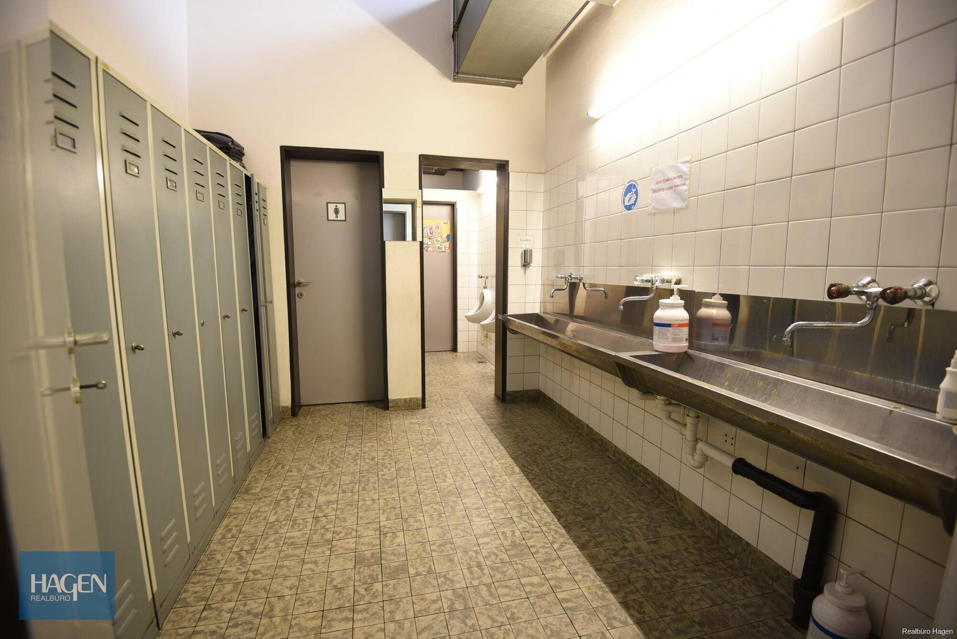 Umkleideraum oder Garderobe mit Spinden, Waschbecken und einem Urinal im Hintergrund, geflieste Wände und Böden.