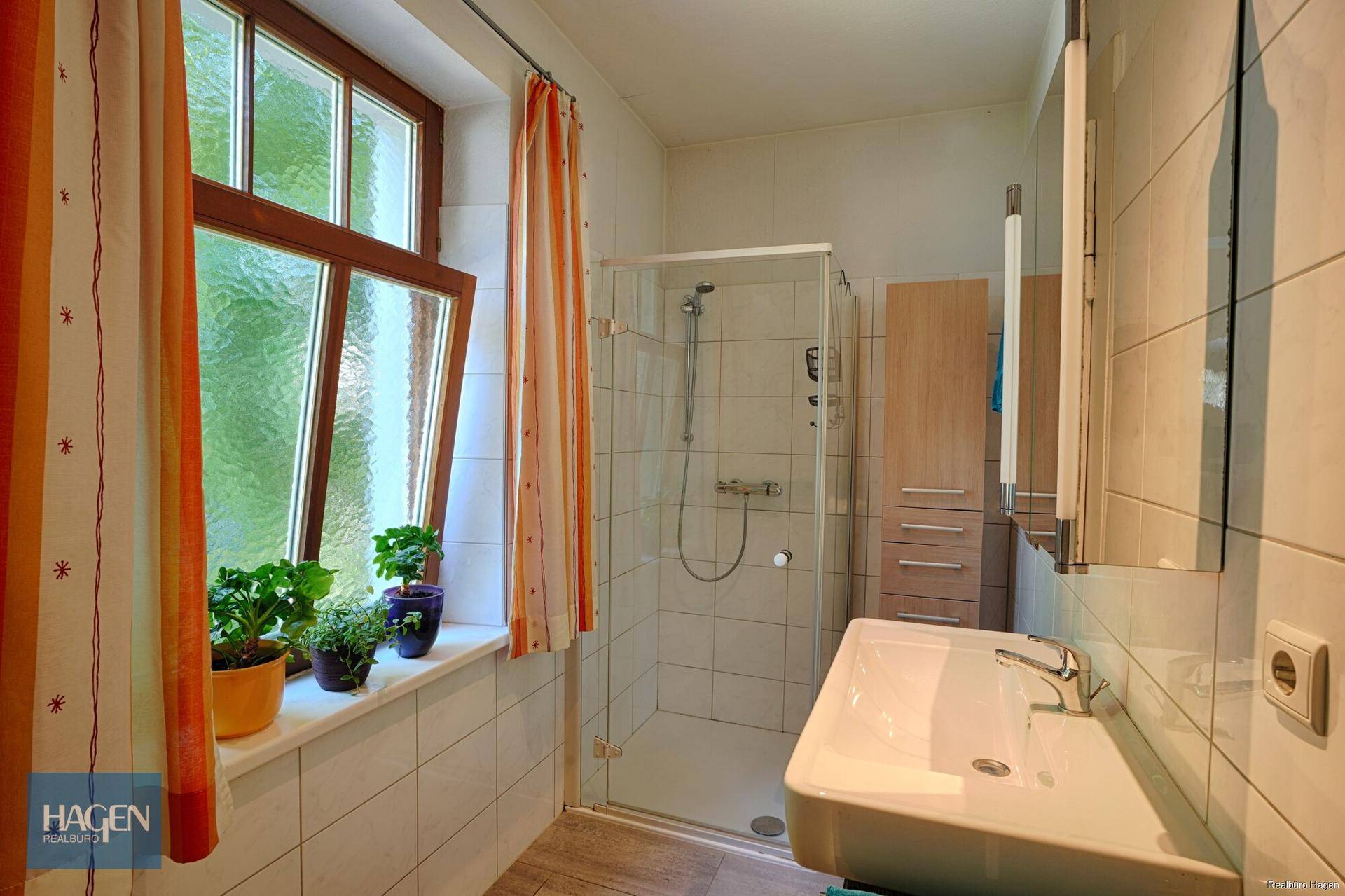 Helles Badezimmer mit Duschkabine, Waschbecken und Fenster mit bunten Vorhängen.