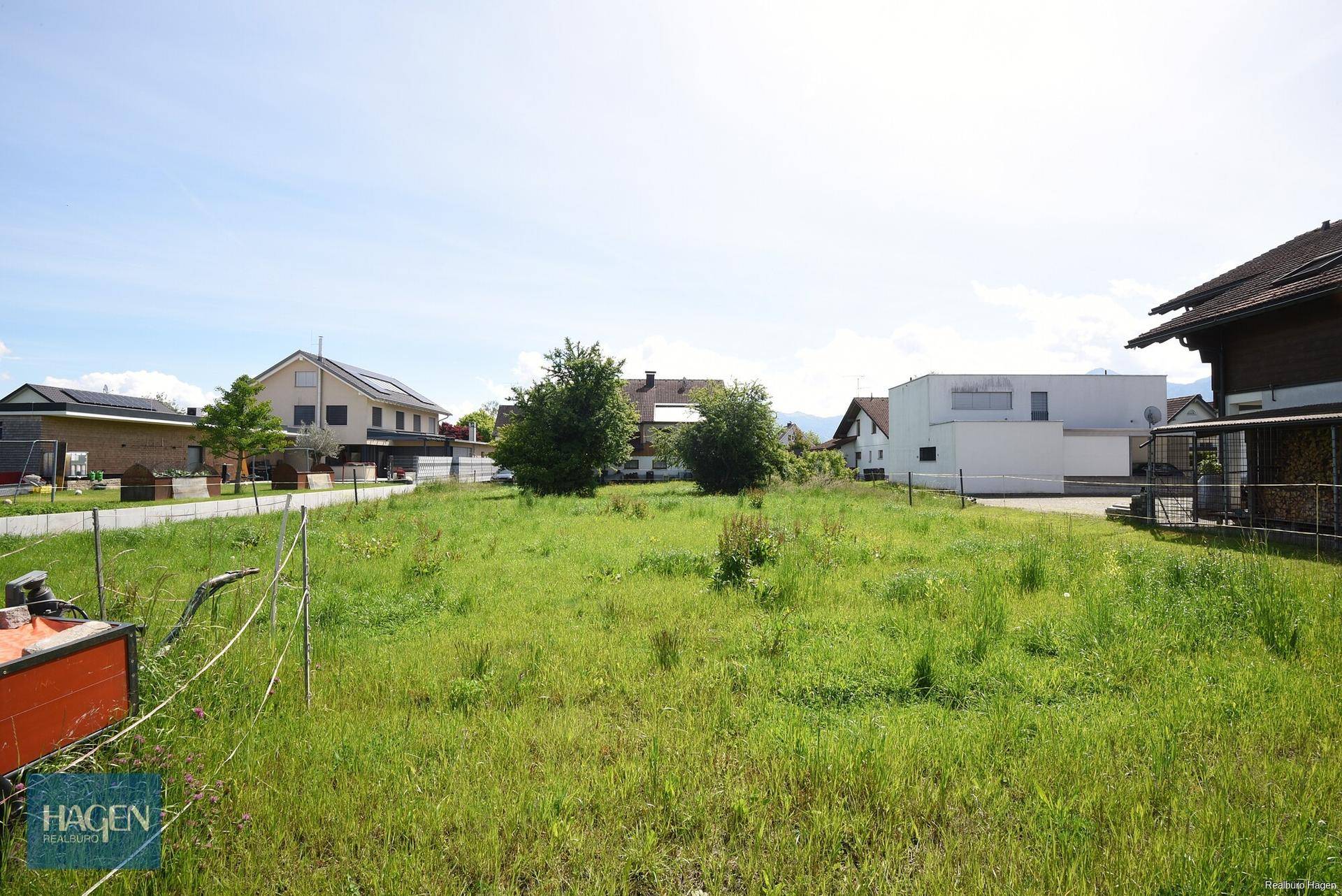 Unbebautes Grundstück mit Wiese und vereinzelten Büschen, umgeben von Wohnhäusern in ländlicher Umgebung.