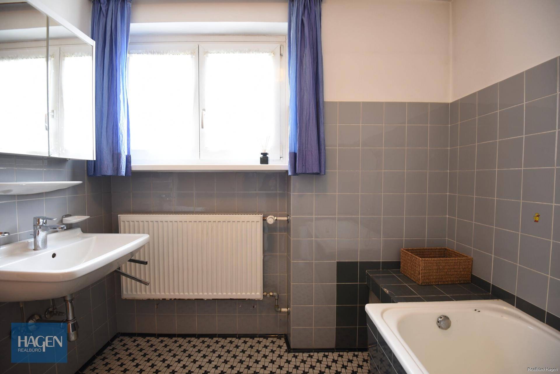 Badezimmer mit grauen Fliesen, Waschbecken, Badewanne und Fenster.