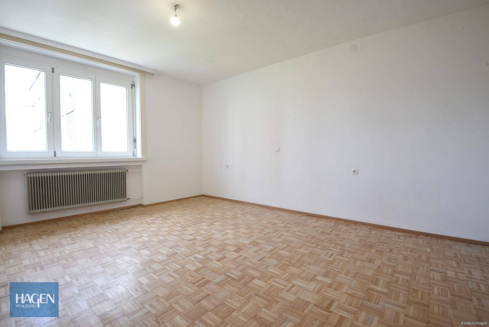 Leeres Zimmer mit Parkettboden und großen Fenstern, die eine helle Atmosphäre schaffen.