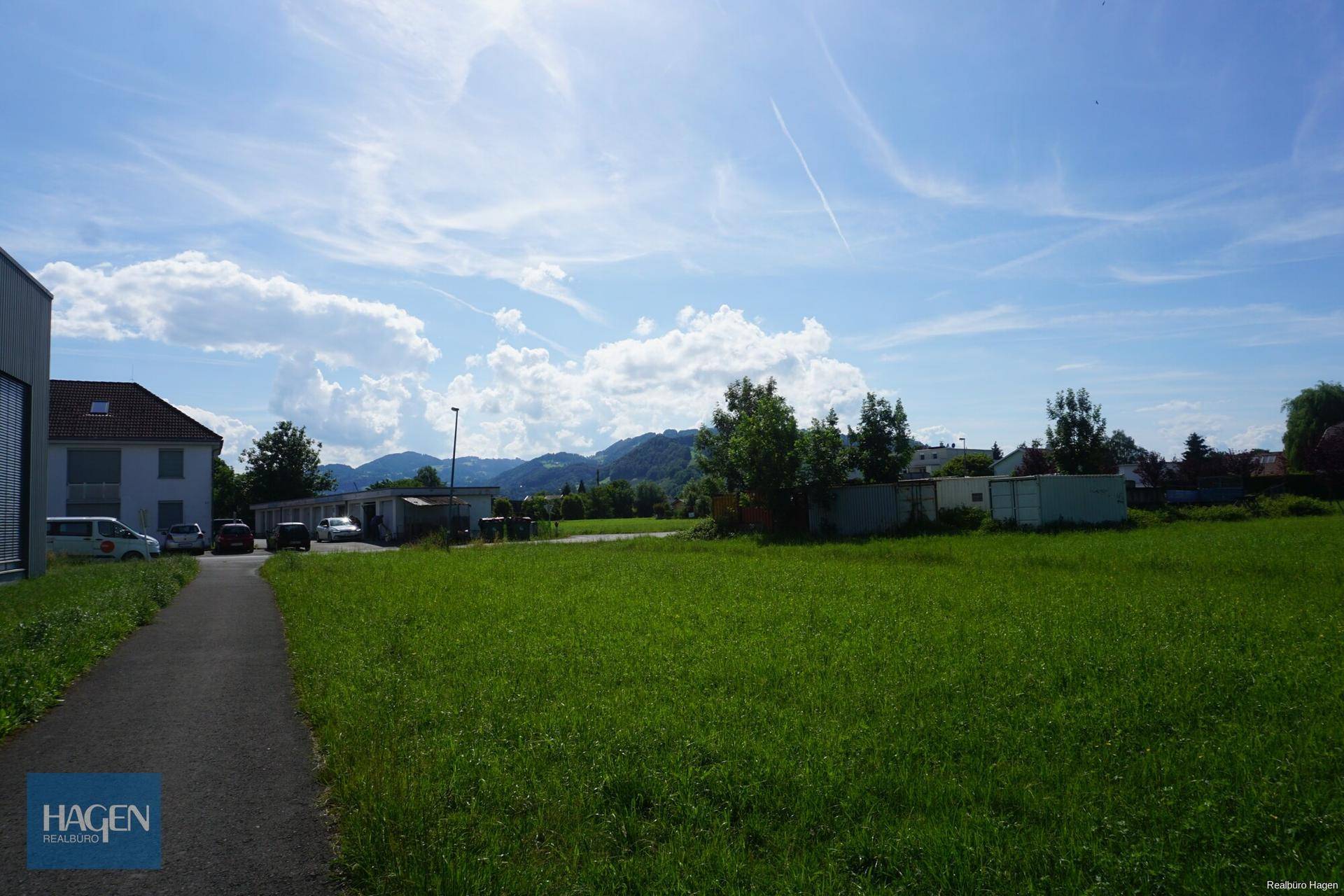 Grüne Wiese mit einem Weg, einem Gebäude und Bergen am Horizont unter blauem Himmel, die eine weite Sicht bietet.