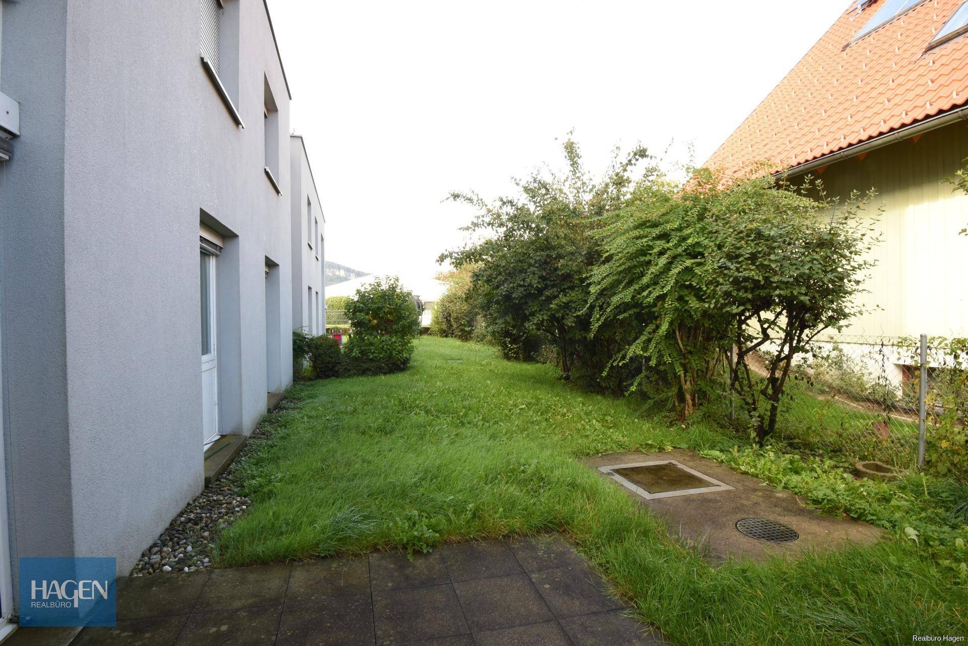 Seitenansicht des Gebäudes mit schmalem Rasenstreifen und dichten Büschen.