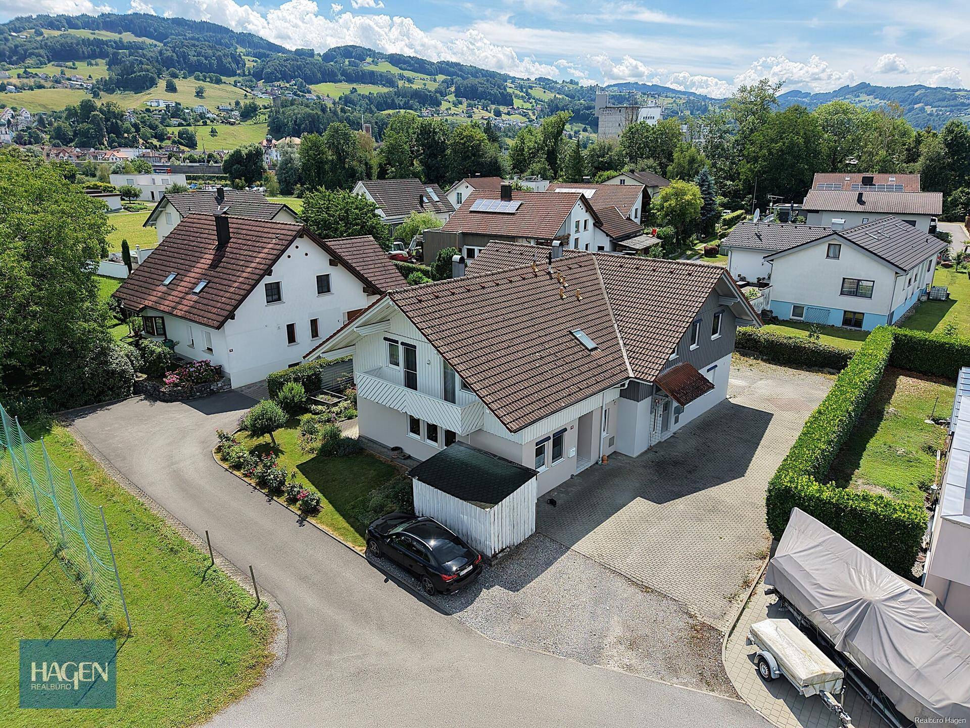 Luftaufnahme des Einfamilienhauses mit umliegender Wohnsiedlung, grüner Landschaft und Bergen im Hintergrund.