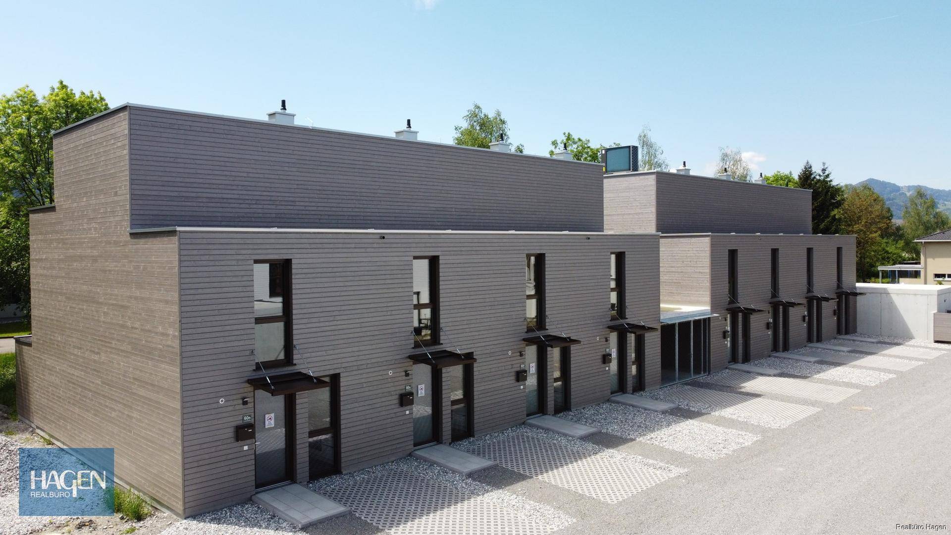 Moderne Reihenhäuser mit grauer Fassade und gepflastertem Vorplatz unter blauem Himmel.