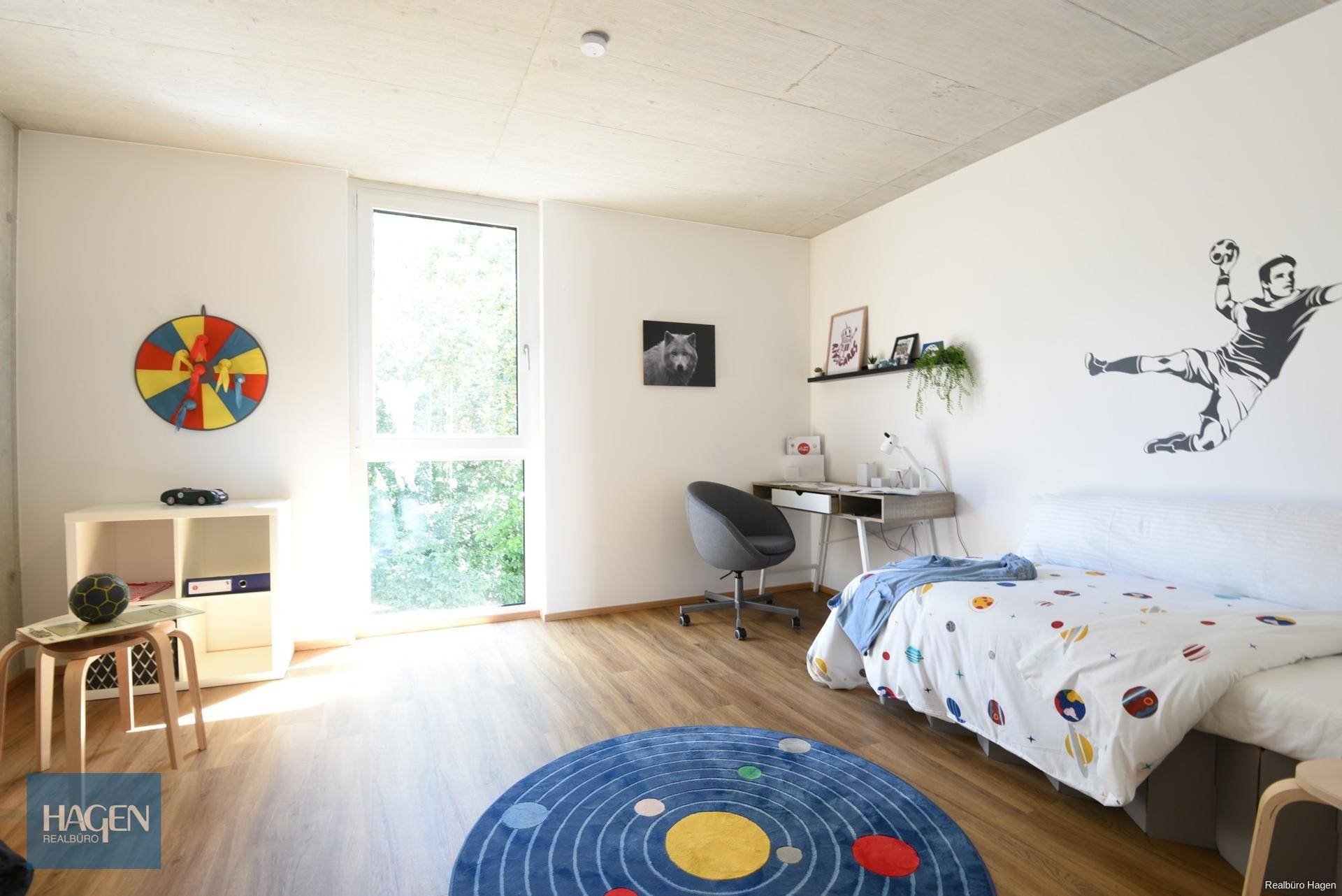 Kinderzimmer mit Holzboden, Schreibtisch, Bett und großem Fenster mit Blick ins Grüne.