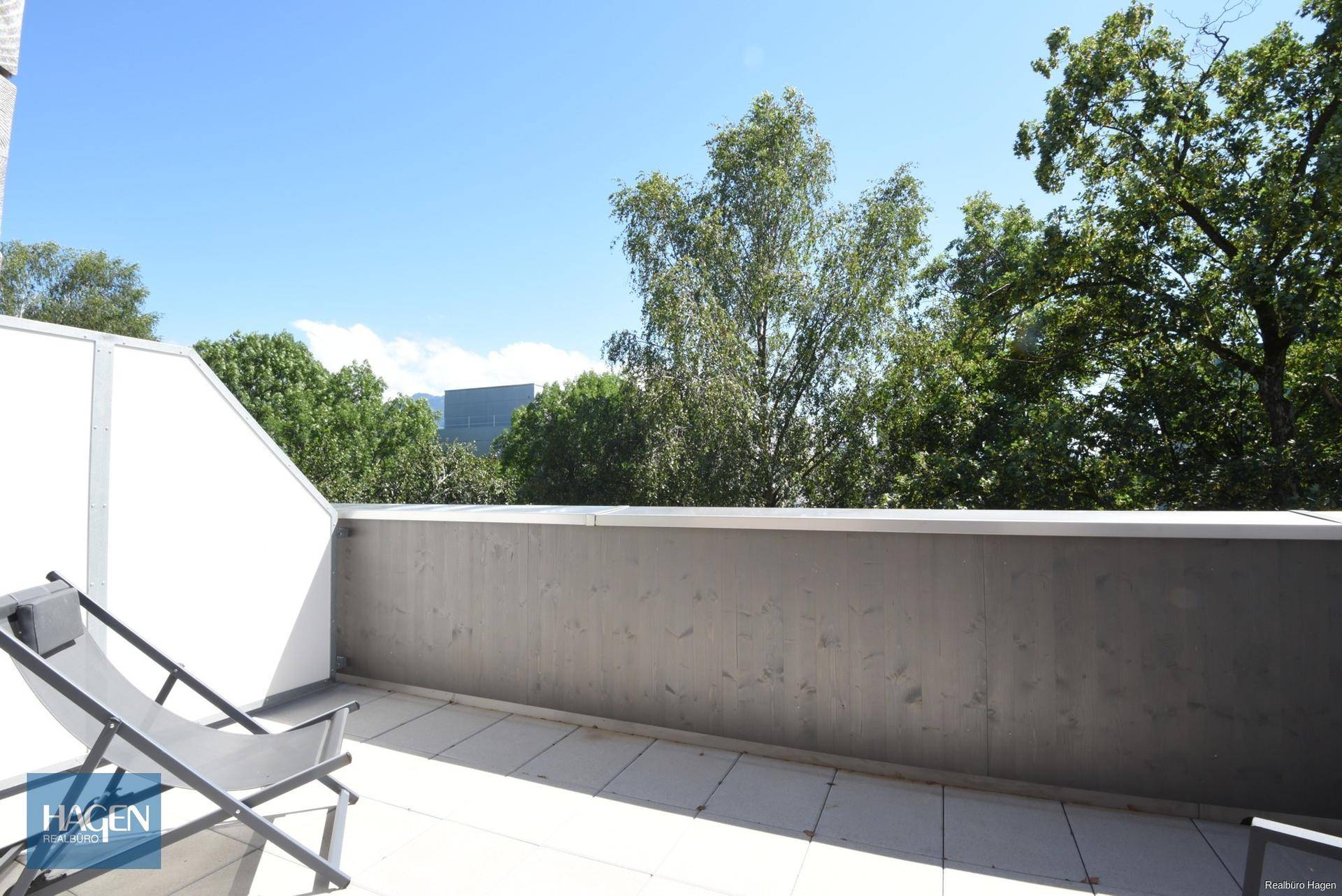 Geräumige Dachterrasse mit Fliesenboden und Liegestuhl, umgeben von Bäumen.
