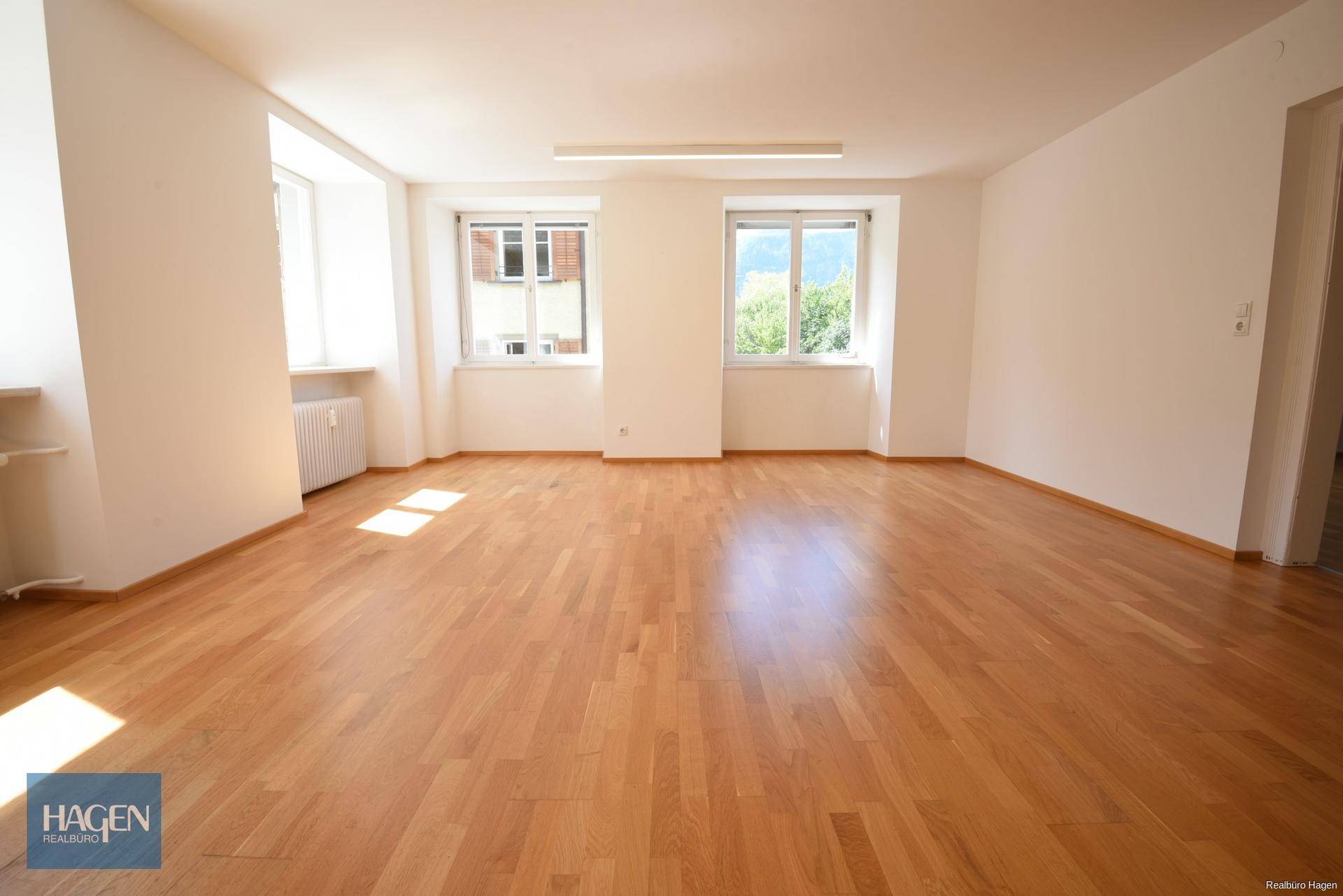 Helles Wohnzimmer mit Parkettboden und zwei großen Fenstern, die viel Tageslicht hereinlassen.