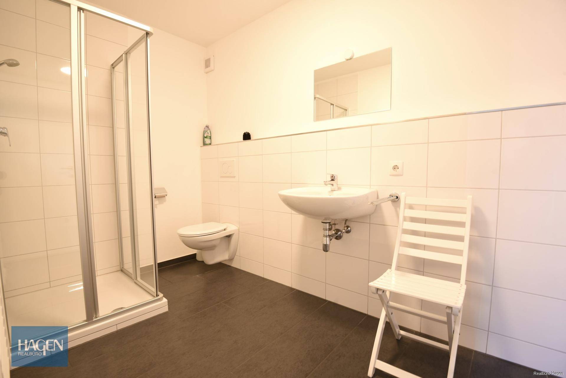 Modernes Badezimmer mit Duschkabine, Waschbecken und wandhängendem WC auf dunklen Fliesen.