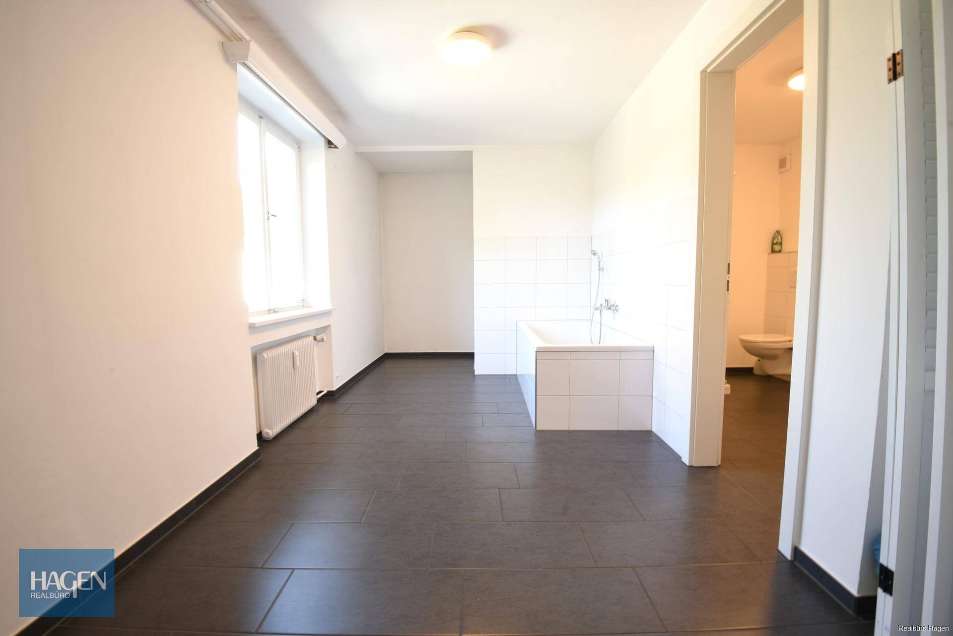 Geräumiges Badezimmer mit dunklen Fliesen, einer Badewanne und Zugang zu einem weiteren Badbereich.