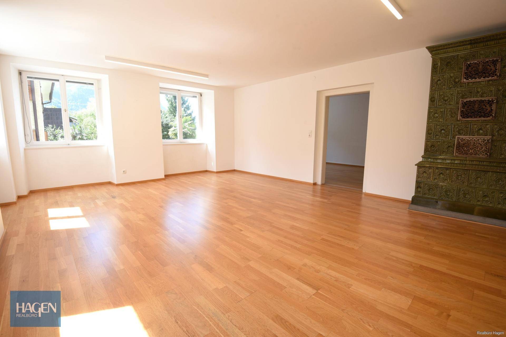 Geräumiges Wohnzimmer mit hellem Parkettboden, großen Fenstern und einem traditionellen Kachelofen.