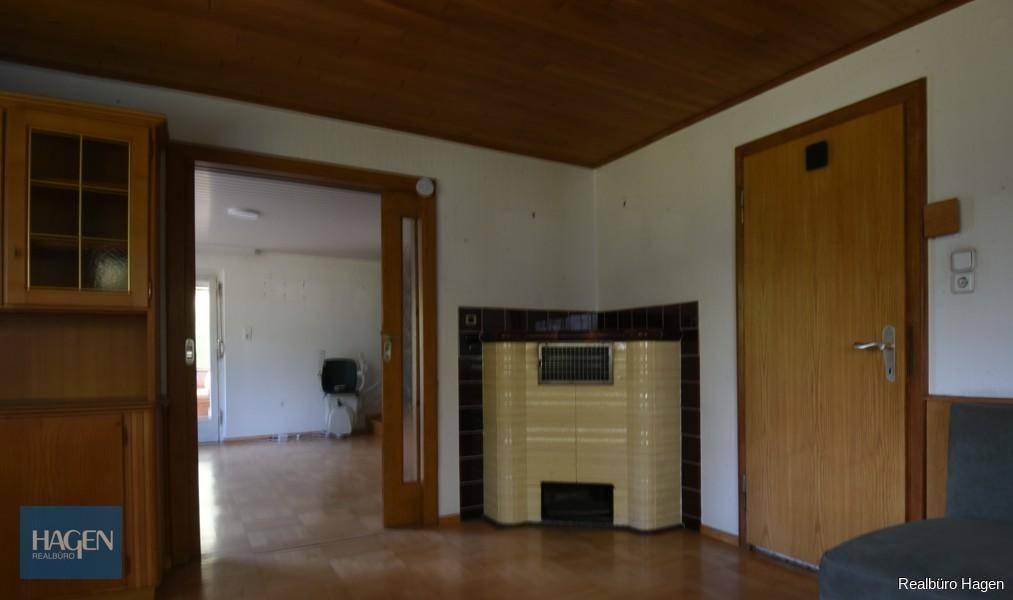 Wohnraum mit traditionellem Kachelofen, Holzboden und Zugang zu einem weiteren Zimmer.