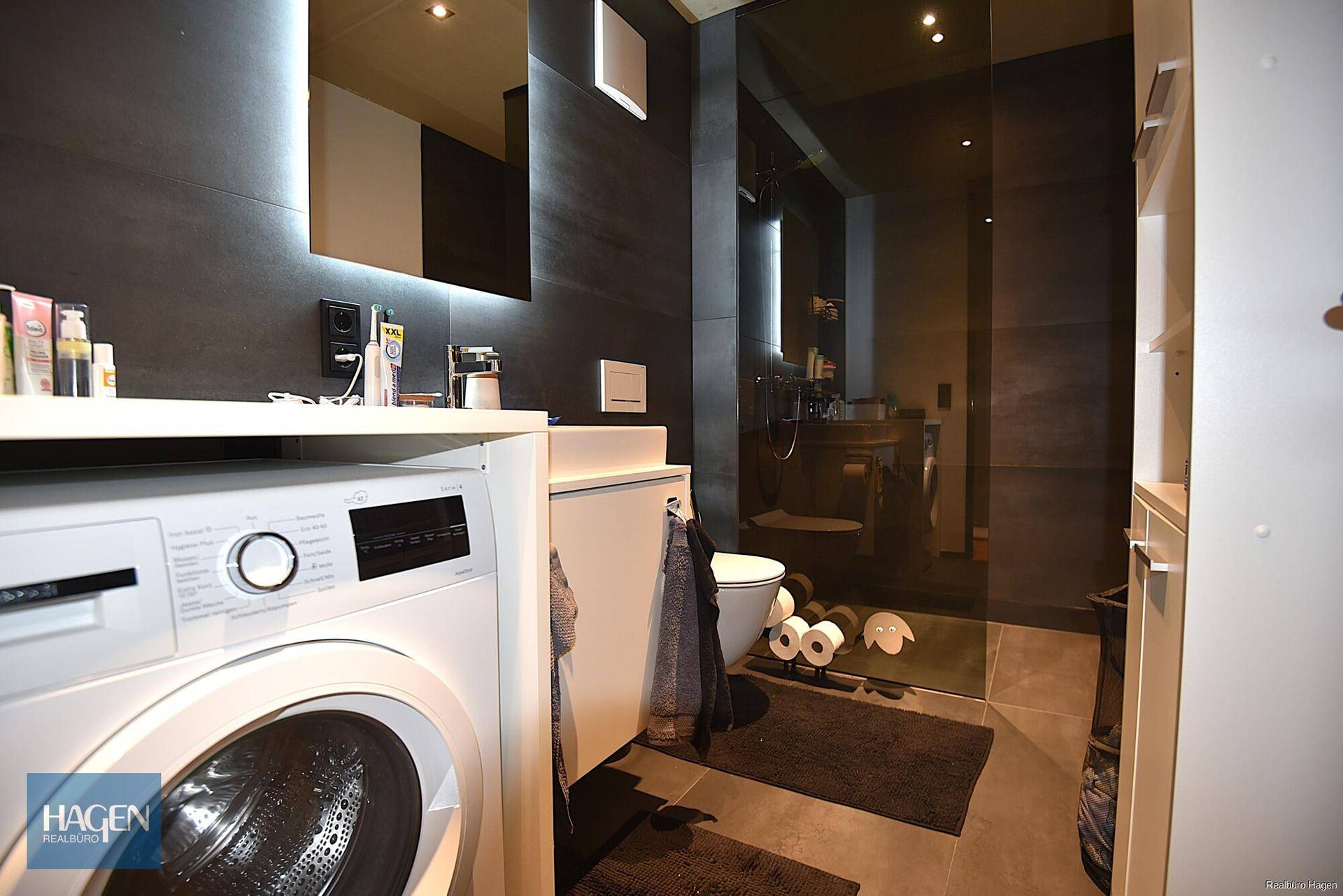 Modernes Badezimmer mit Waschmaschine, Toilette und begehbarer Dusche in dunklen Fliesen.