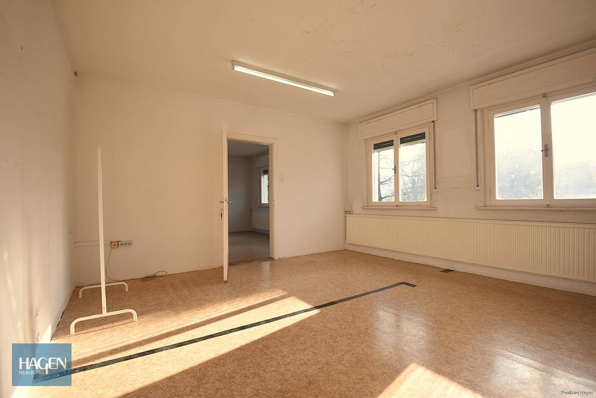 Heller Büroraum mit Fenstern, Heizkörpern und Linoleumboden.
