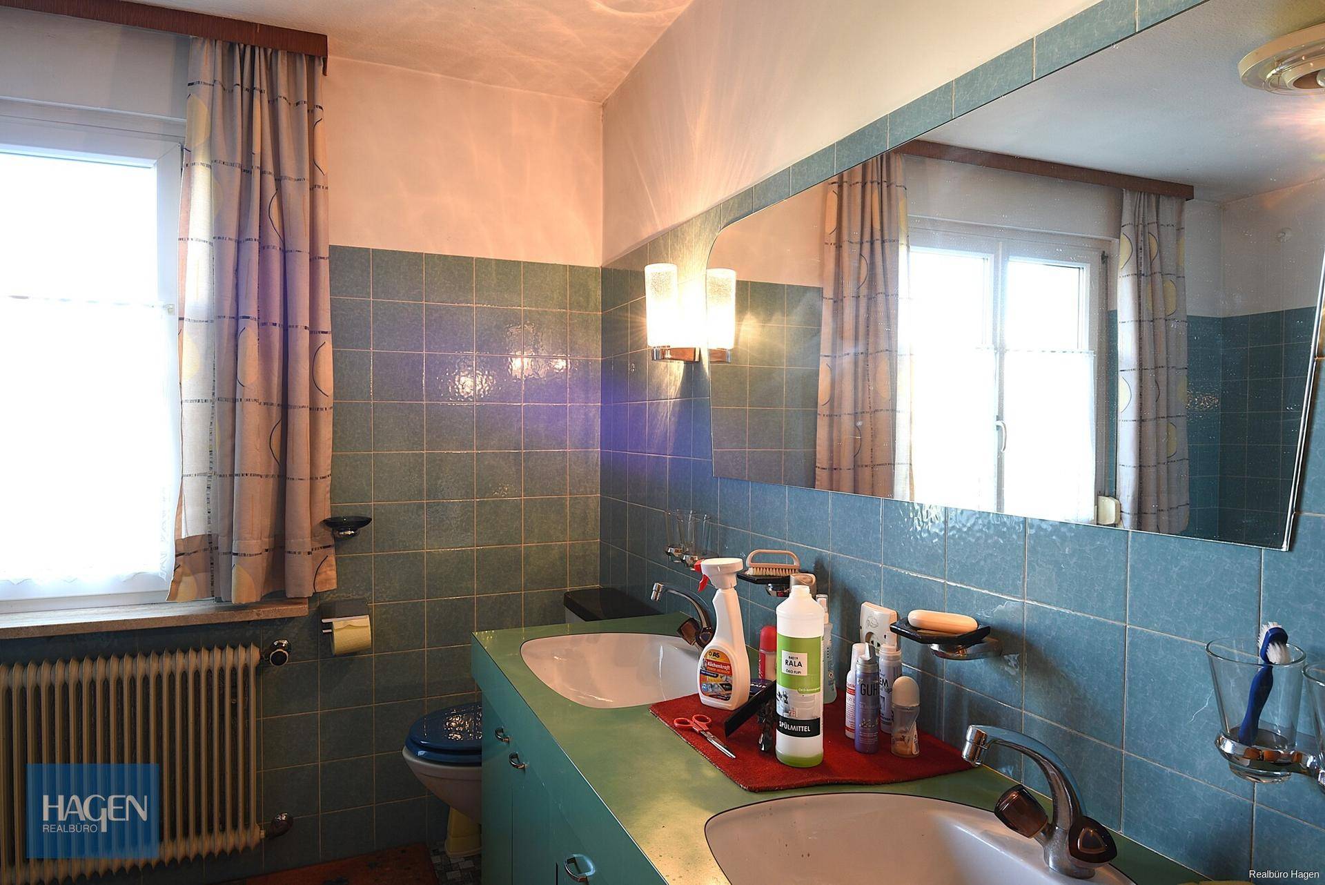 Badezimmer mit zwei Waschbecken, großem Spiegel und blauen Fliesen im 80er-Jahre-Stil.