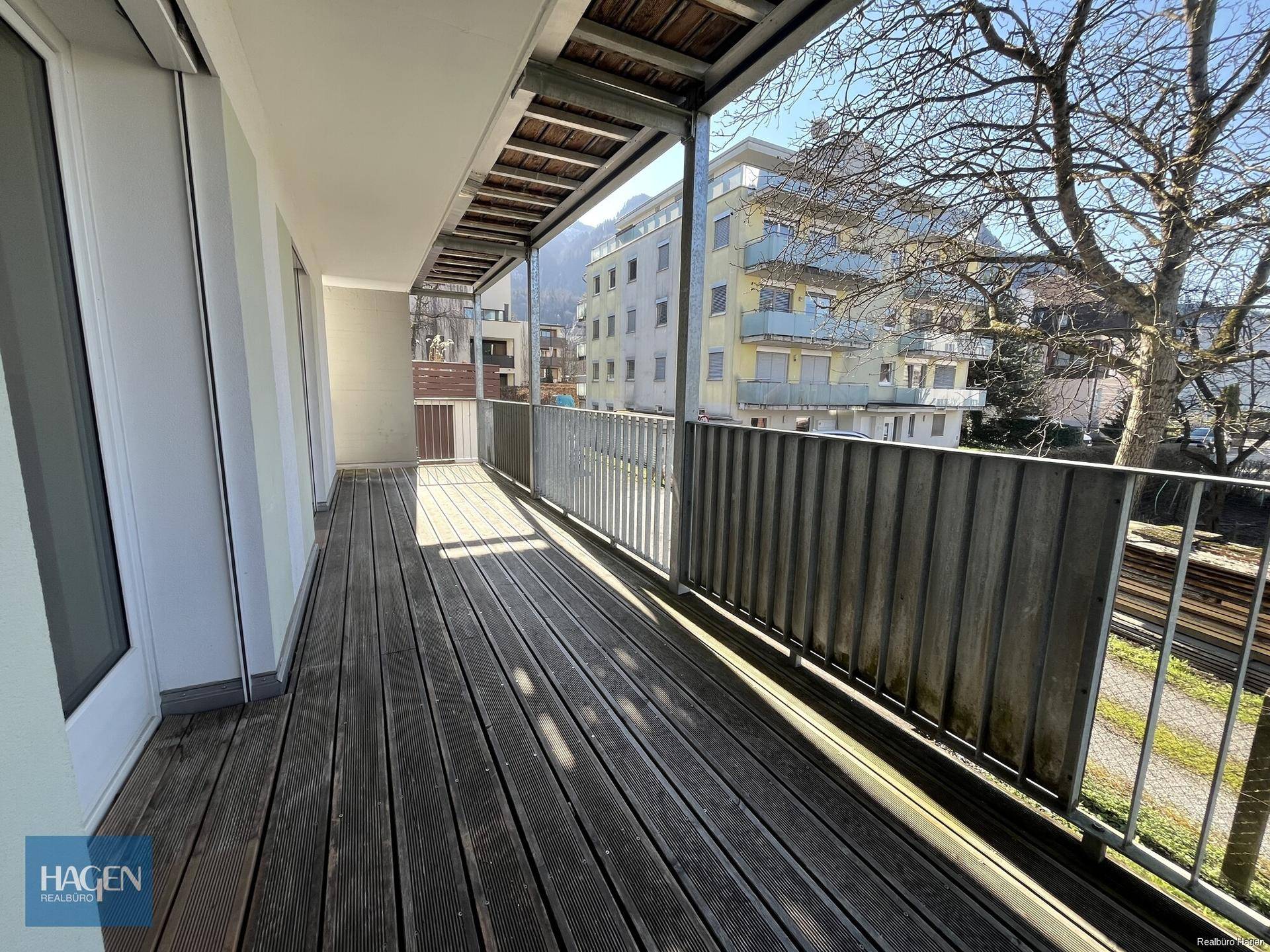 Sonniger Balkon mit Holzboden und Blick auf die umliegende Wohnbebauung und Bäume.