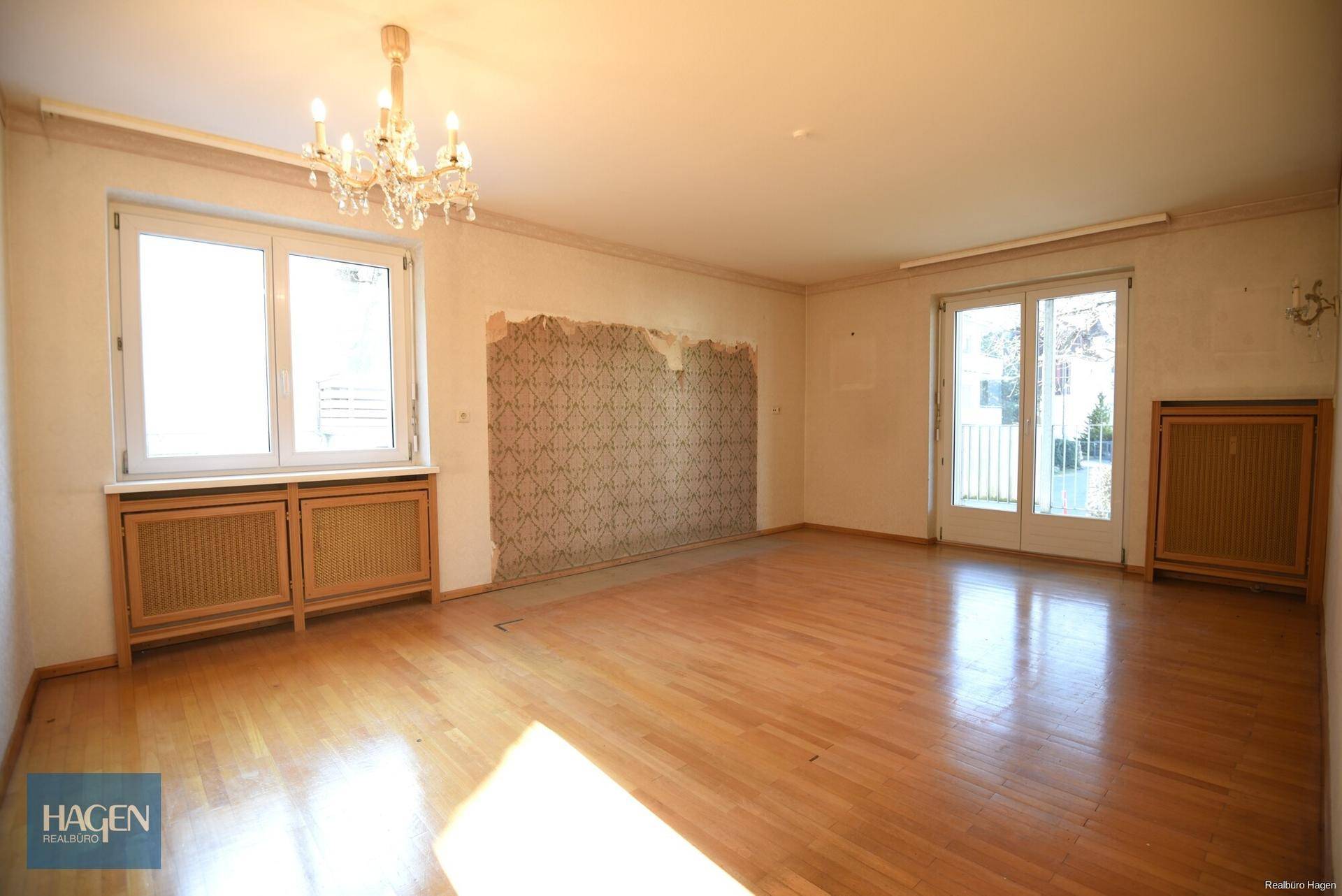 Geräumiges Wohnzimmer mit Laminatboden, Kronleuchter und Zugang zum Balkon, teilweise renovierungsbedürftig.