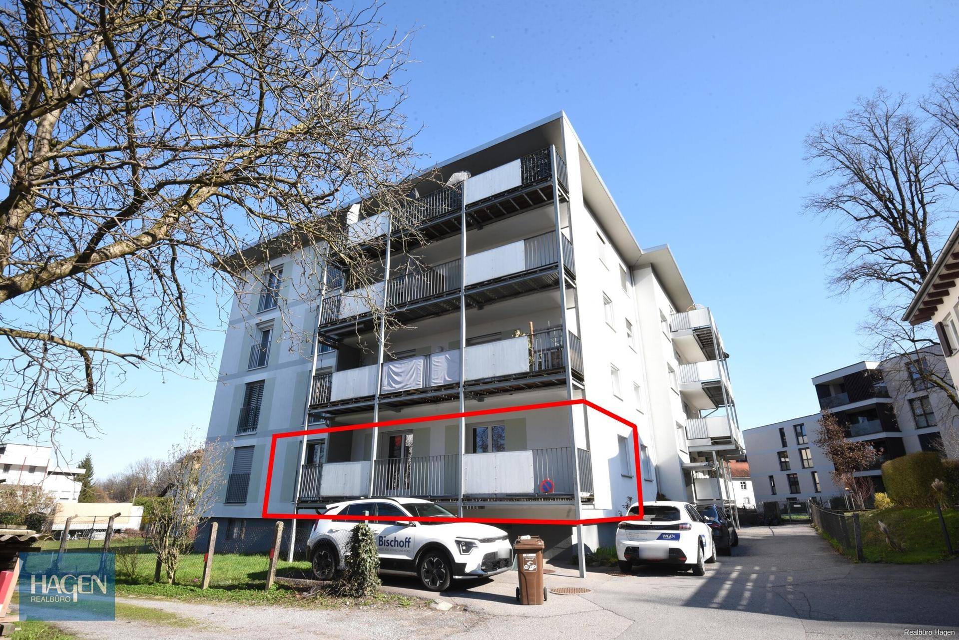 Außenansicht eines Mehrfamilienhauses mit Balkonen und geparkten Fahrzeugen davor.