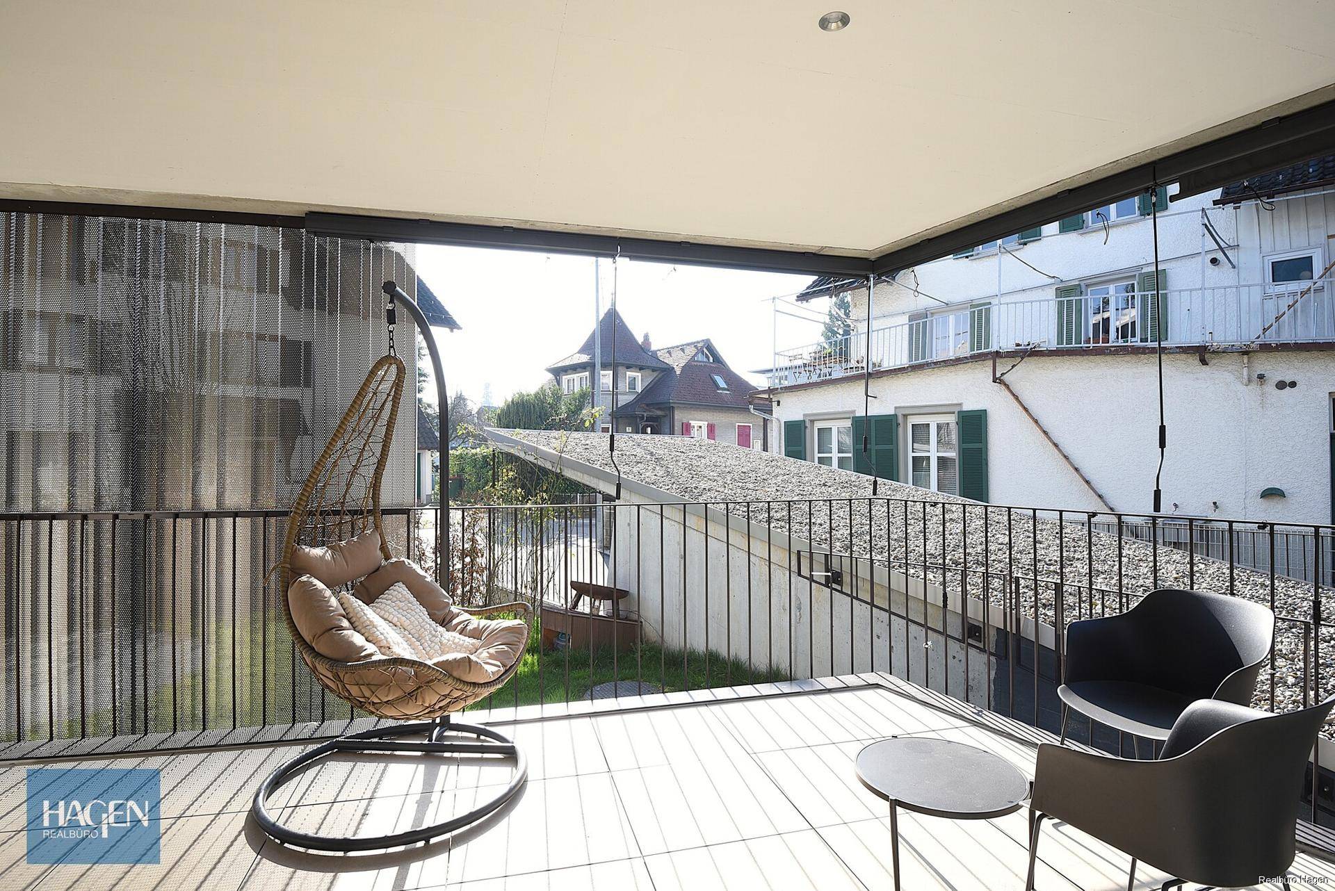 Heller Balkon mit Holzboden, Hängesessel und Blick auf die Umgebung.