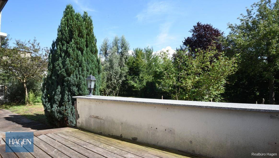 Holzterrasse mit Steinmauer und Blick ins Grüne, umgeben von hohen Bäumen.