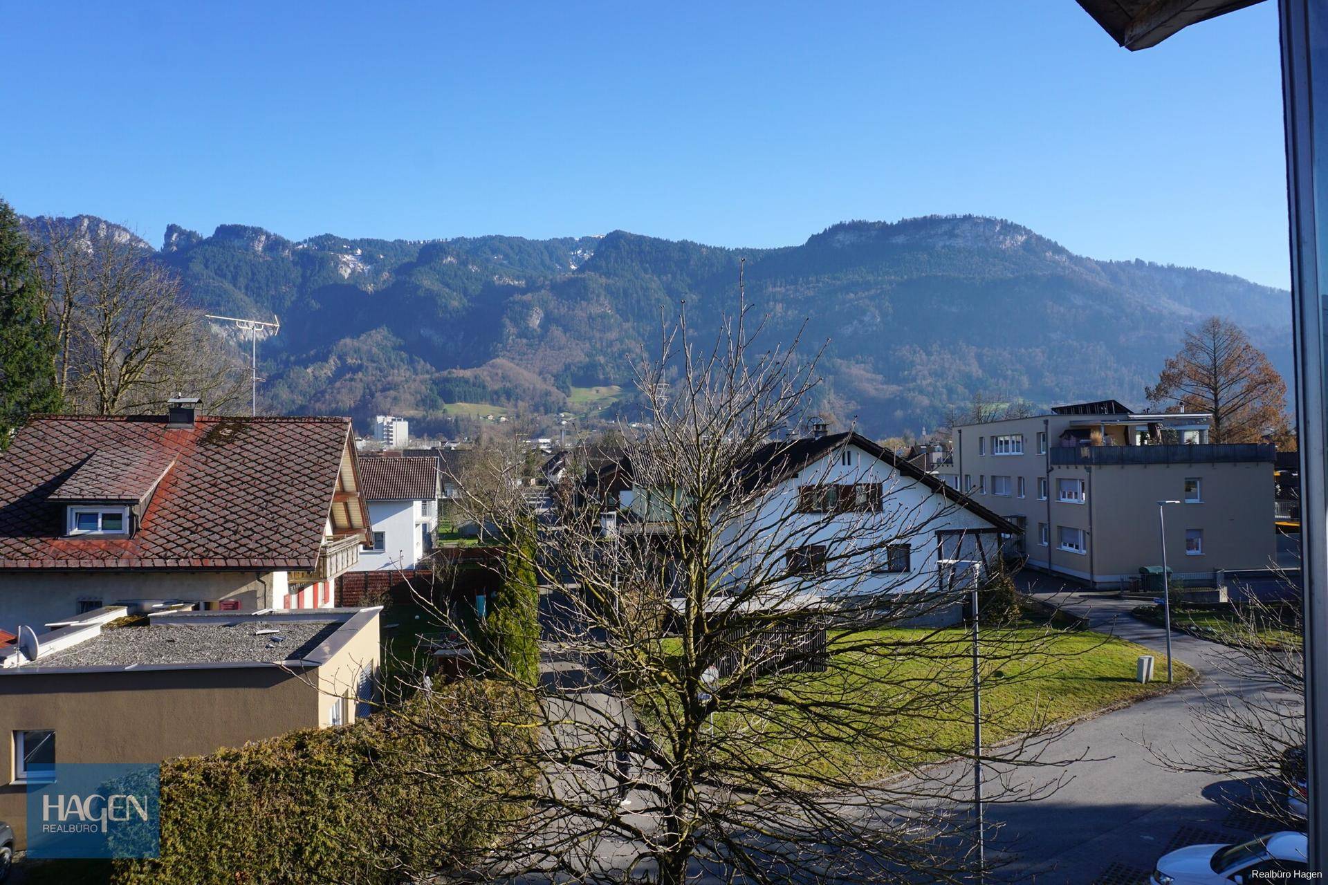 Weitblick über eine Wohnsiedlung mit Häusern und majestätischen Bergen im Hintergrund unter blauem Himmel.