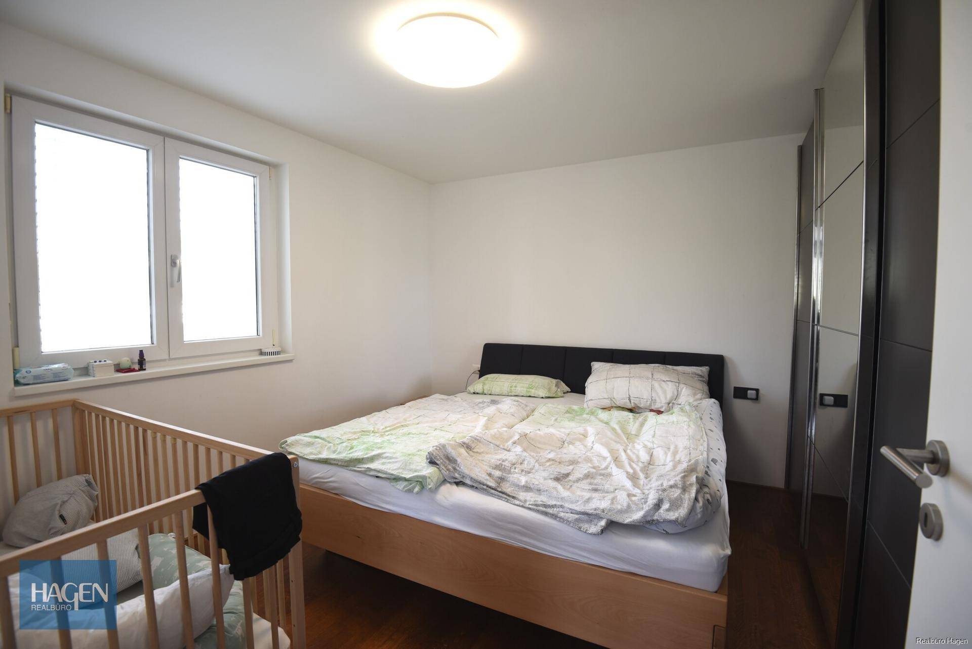 Schlafzimmer mit Doppelbett, Kinderbett und großem Fenster, das viel Licht hereinlässt.