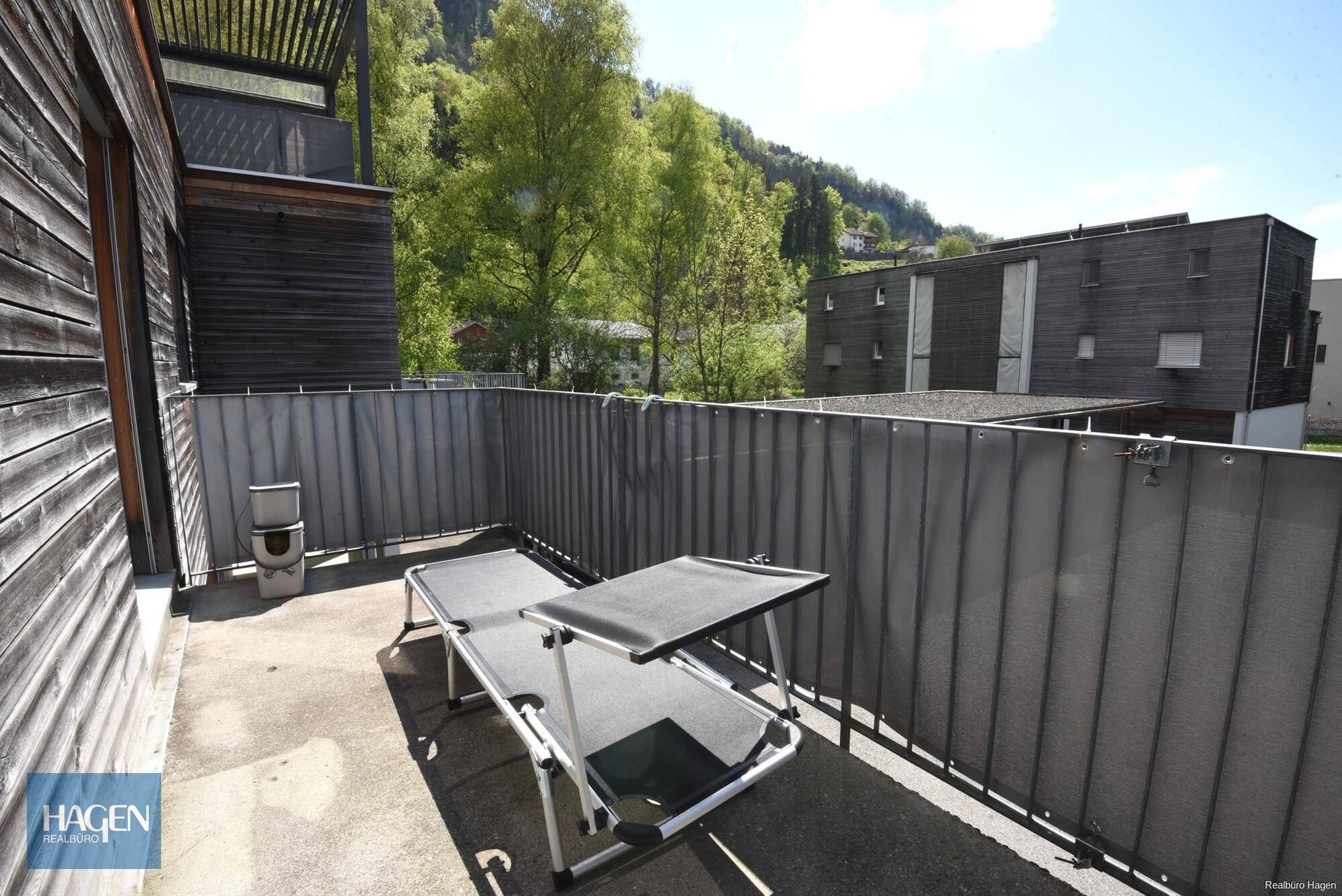 Sonniger Balkon mit Liegestuhl und Sichtschutz, umgeben von grüner Landschaft.