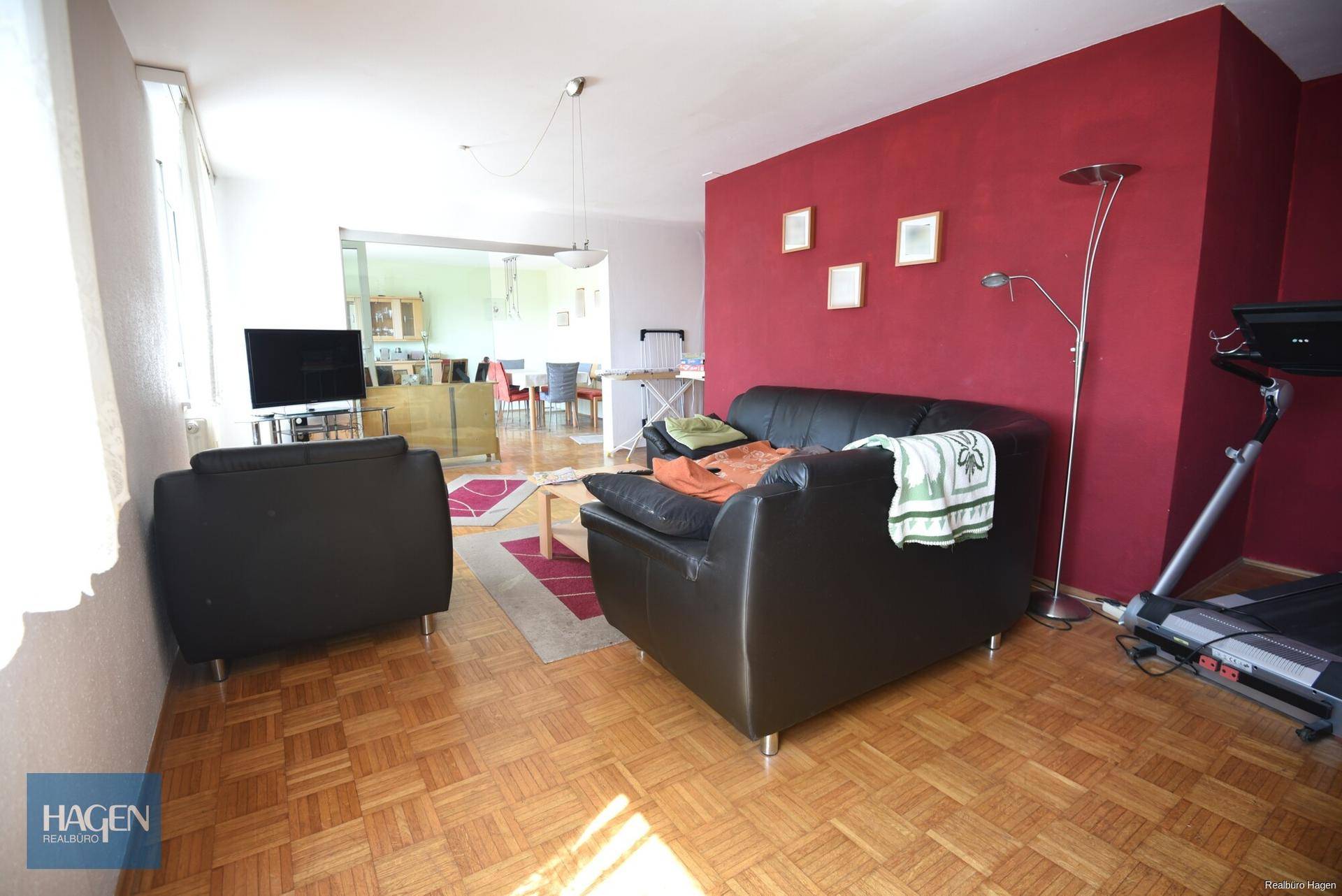 Helles Wohnzimmer mit Parkettboden, roter Akzentwand und offenem Zugang zum Essbereich.